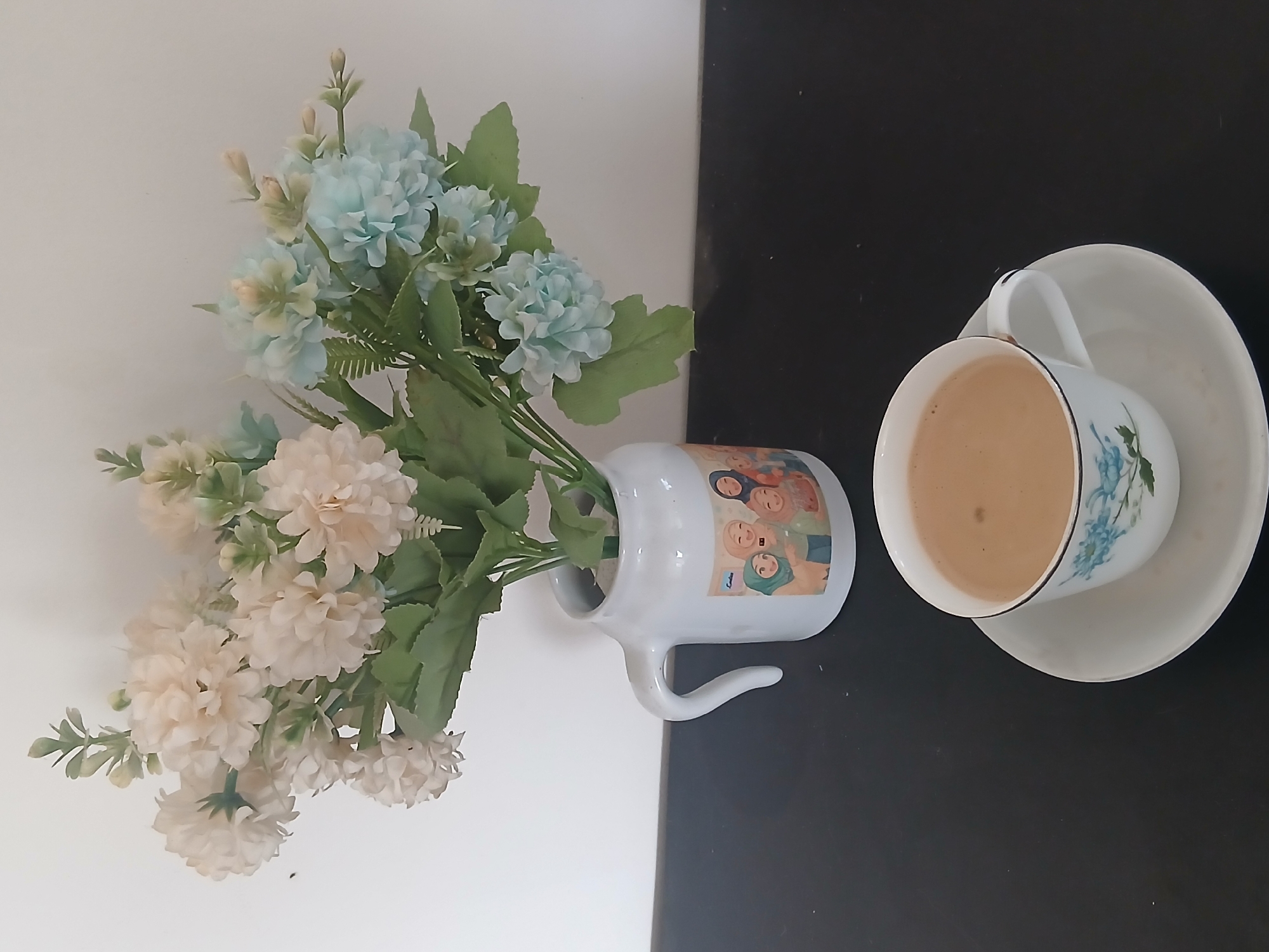 A cup of coffee sits on a saucer on a dark surface, with a bouquet of artificial flowers in a white vase behind it. The vase is decorated with a cartoon image of several women wearing hijabs. The background is a plain white wall. There is no indication of the location in West Jakarta, Indonesia.