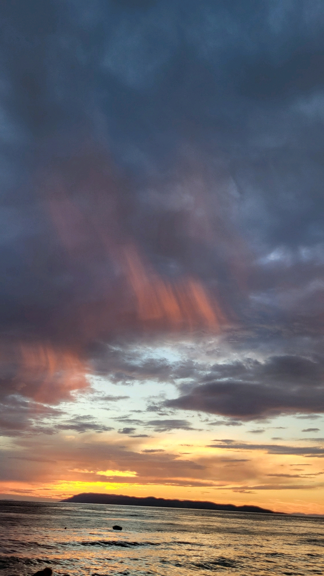 The image depicts a dramatic sunset over the Adriatic Sea, viewed from the coast of Općina Tučepi, Croatia. Dark, moody clouds dominate the upper portion of the frame, with streaks of pink and orange light filtering through them, creating a textured, almost painterly effect. Below the clouds, a vibrant band of yellow and orange light illuminates the horizon, where a silhouetted mountain range stretches across the scene. The sea in the foreground is choppy, with light reflecting off the waves, and two small objects, possibly buoys or small boats, are visible in the water. The overall impression is one of natural grandeur and a fleeting, powerful display of light and color.
