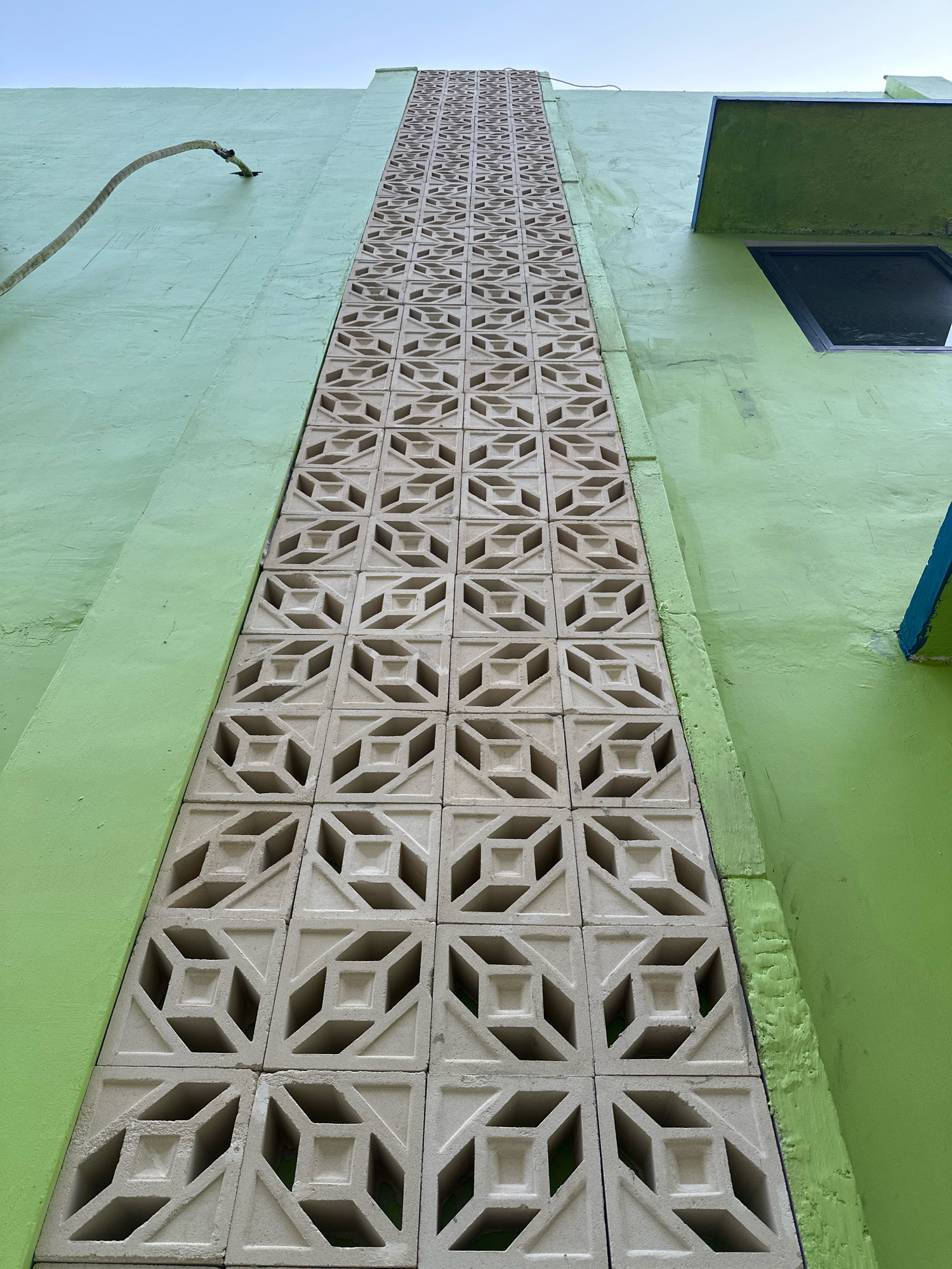An upward-facing, wide-angle shot captures a section of a light green building in Saruni, Indonesia. Dominating the center is a vertical column of cream-colored decorative concrete blocks, each featuring an intricate geometric pattern of interlocking squares and triangles. The blocks are arranged in a repeating, precise grid. To the left of the concrete blocks, the building's smooth, light green wall extends upwards, with a curved, brown cable attached to it and arching away from the wall. To the right of the blocks, the green wall continues, with a dark-framed window visible towards the top. A section of a brighter blue painted edge is also visible on the far right, just above the window. The sky above is a pale, overcast blue. The overall scene is one of architectural detail on an exterior wall, with a focus on the textural contrast between the smooth painted surface and the patterned concrete.