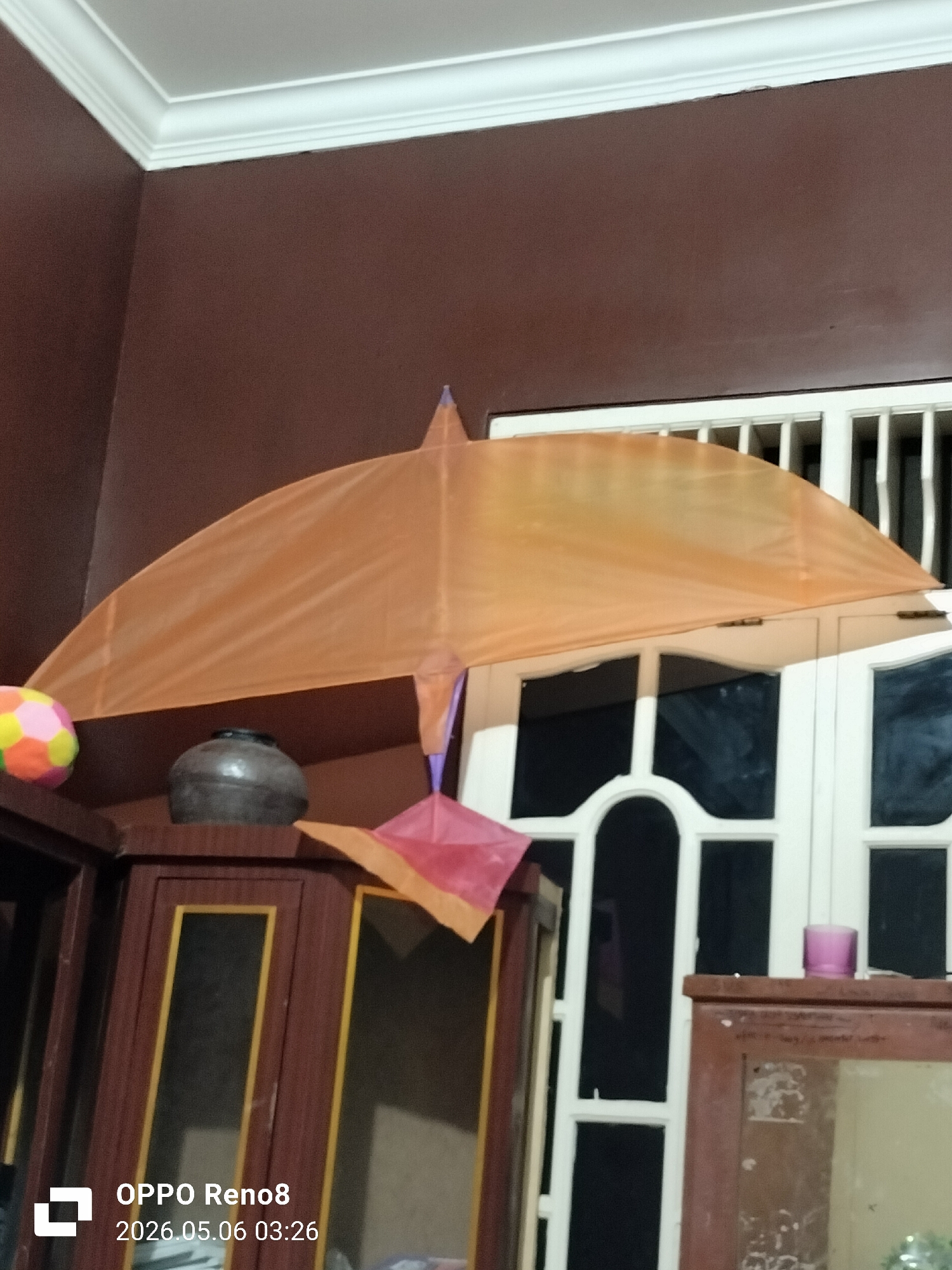 The image shows an indoor scene with a large orange kite in the foreground. The kite is positioned in front of a white window with vertical and arched panels. To the left of the kite is a wooden cabinet with glass panels and brown framing. On top of the cabinet, there is a dark-colored pot and a multi-colored ball. The walls are brown, and the ceiling has white molding. A small purple object sits on a wooden surface to the right of the kite. The image metadata indicates that it was taken with an OPPO Reno8 on May 6, 2026, at 03:26. The location is not explicitly stated within the image but given the prompt, it is assumed to be in Lhokseumawe, Indonesia. No people are visible.