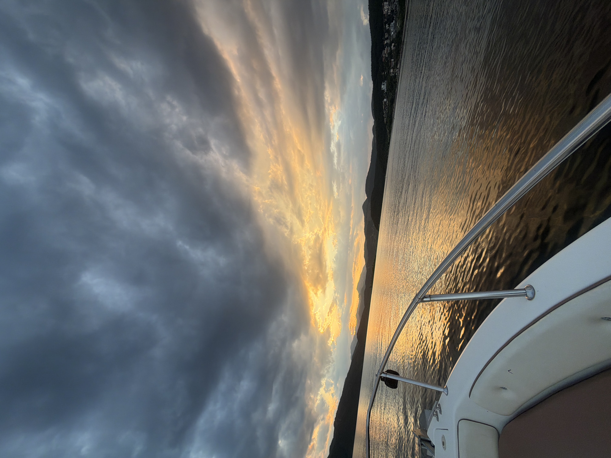 A boat's perspective of a lake at sunset, with the city of Glavatartsi, Bulgaria visible on the shore. The sky is filled with dark, dramatic clouds with pockets of golden light breaking through. The lake's surface reflects the sunset, creating a shimmering, warm glow. The foreground shows a portion of the white boat, including a stainless steel railing and beige seating. The distant shore is lined with dark green hills and a scattering of buildings.