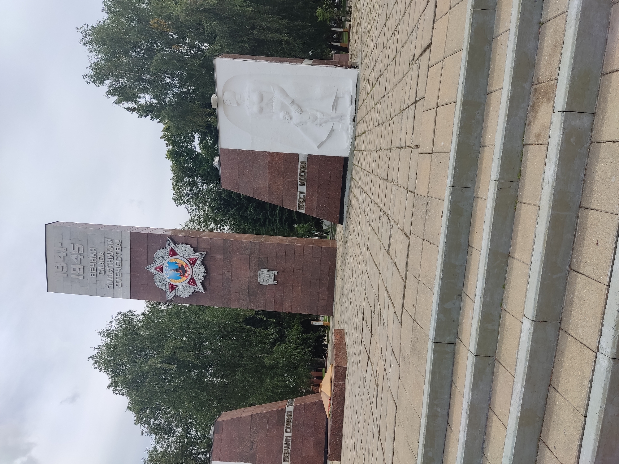 The image captures a war memorial in the city of Noginsk, Russia. The central monument is a tall, granite obelisk inscribed with "1941-1945" and "ETERNAL GLORY TO THE DEFENDERS OF THE FATHERLAND!" Below the inscription is a large, red star with a medal at its center featuring the hammer and sickle emblem. To the left of the obelisk is a smaller, reddish-brown monument with text that appears to read "BERLIN SOFIA." In front of this monument is a flame of remembrance. To the right of the obelisk is another monument with a reddish-brown angled surface inscribed with "BREST MOSCOW," and a large white relief sculpture depicting a woman holding a child. The memorial is situated on a paved plaza made of square tiles. Steps lead up to the plaza from the foreground. Trees line the background, and the sky is overcast. No people are visible in the immediate vicinity of the monuments.