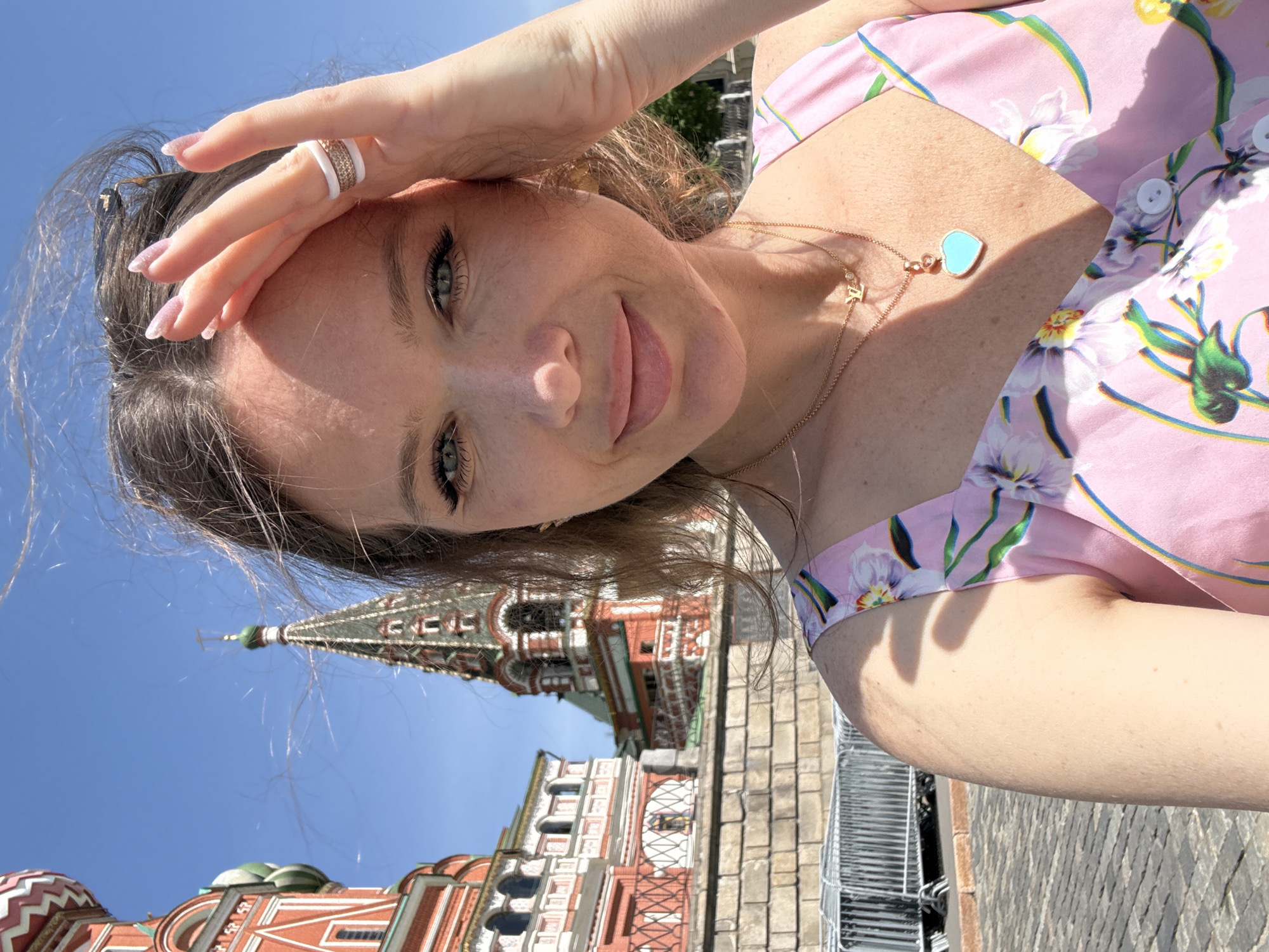 A woman takes a selfie in front of Saint Basil's Cathedral in Moscow, Russia. The woman has light skin, long brown hair, and is wearing a pink floral dress. She is holding her hand to her forehead, and has rings on her fingers. The Cathedral is visible in the background, with its colorful domes and intricate details. The sky is blue and clear. The scene is outdoors, and is shot during the day.