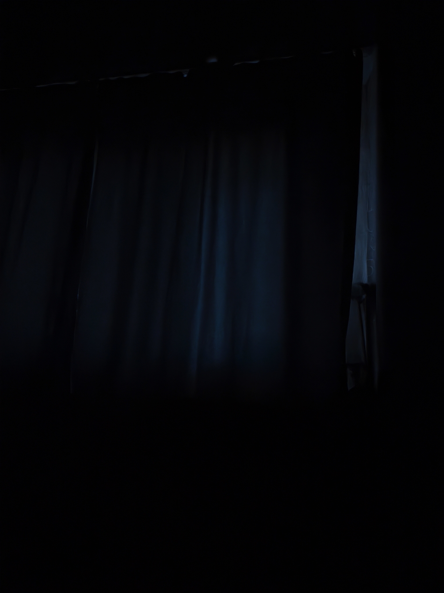 The image depicts a dimly lit interior with dark blue curtains. A faint light source is visible on the right side, possibly from behind another set of sheer curtains. The overall impression is one of darkness and obscured visibility. There are no people or specific objects clearly identifiable, and the environment appears to be a room. The image does not provide any information about the city of Leninsk-Kuznetsky, Russia.