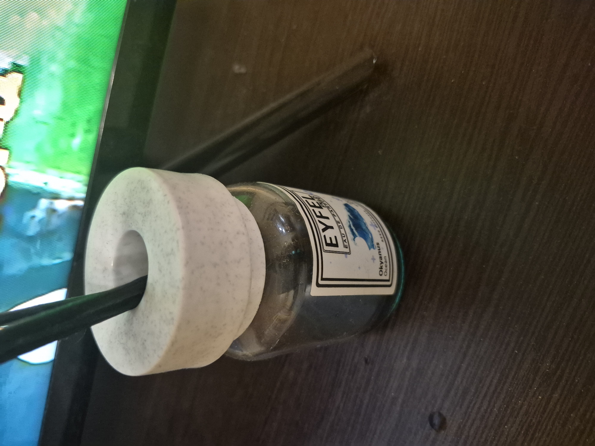 An indoor scene, likely a desk or table in Yerevan, Armenia, is depicted from an oblique, slightly elevated angle. The primary subjects are a glass bottle, a white cylindrical object, and a black cable, set against a dark wooden surface.
A clear glass bottle lies horizontally, with a white and black label facing upwards. The label clearly shows the brand name "EYFEL" in large letters, followed by "EAU DE PARFUM." Below this, "Okyanus" and "Ocean" are visible, indicating the product's fragrance or type. The bottle contains a dark liquid, possibly blue or green, consistent with an "Ocean" theme.
Partially obscuring the bottle is a textured, white, cylindrical object with a central hole, which appears to be a cable organizer or a counterweight. A black cable passes through this hole, extending towards the left of the frame. To the far left, an electronic screen stands vertically, displaying vibrant, pixelated green and blue imagery, likely from a digital application or video game, with visible horizontal scan lines. A slender, dark rod or cable also rests on the wooden surface behind the bottle and the white cylinder. The wooden surface has a visible grain and appears slightly dusty. No specific activity is taking place; the scene shows a static arrangement of objects. The time of day is indeterminate, but the setting is indoors.