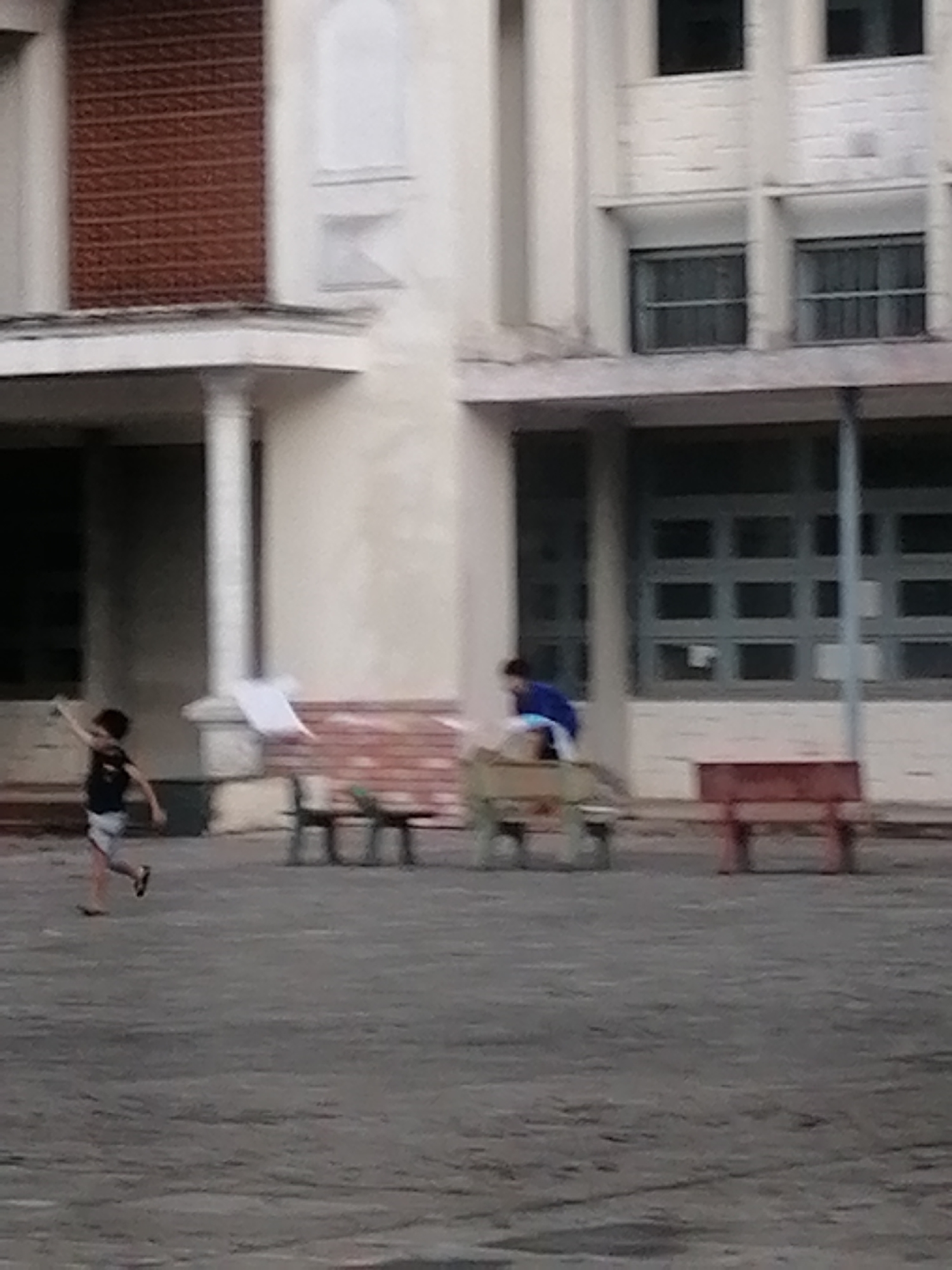 The image shows an exterior scene in Da Lat, Vietnam, in front of a light-colored, multi-story building with a combination of brick and plastered facade. The building has a covered entrance supported by columns. Several window units are visible. In the foreground, two children are present. One child, wearing a dark shirt and light shorts, is running and appears to be throwing a piece of paper. Another child, wearing a blue shirt, is standing on a wooden bench. Several benches are arranged in front of the building. The ground is paved with a gray material. The overall image quality is somewhat blurry.