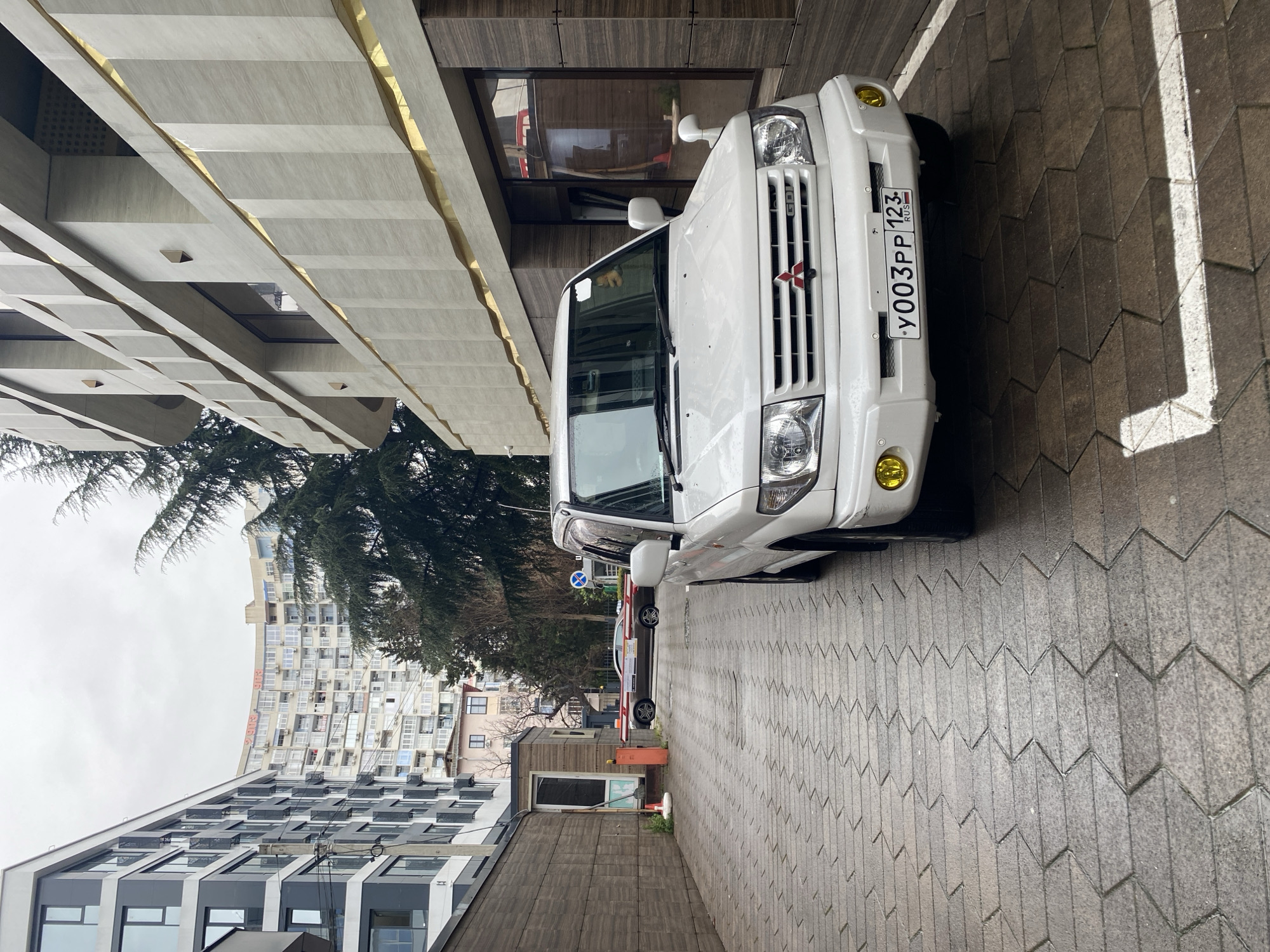 A white Mitsubishi Delica with a Russian license plate "У003РР 123" is parked on a herringbone-patterned concrete brick surface. The vehicle is adjacent to a modern building with gold-accented concrete sections. In the background, there is a taller residential building with numerous balconies. The location is Sochi, Russia.