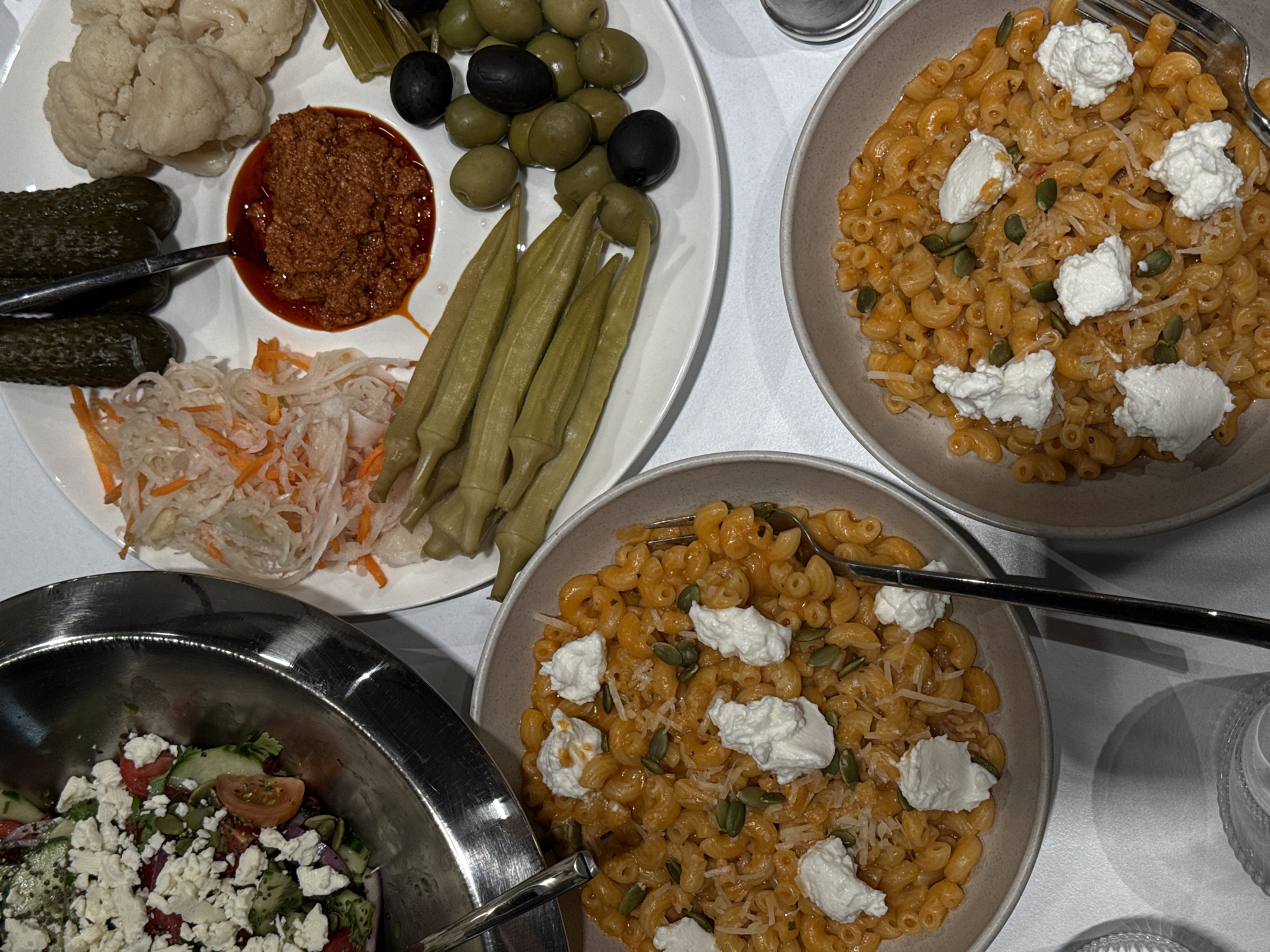 An overhead view reveals a dining table set with multiple dishes on a white tablecloth in Yerevan, Armenia. In the upper left quadrant, a large, polished metallic bowl holds a salad comprising diced cucumbers, tomato wedges, red onion, green leaves, and dark seeds, all topped with substantial crumbles of white feta cheese. A metallic utensil is partially visible within the salad bowl.
To the right, a large white oval platter is laden with various pickled and preserved items. These include a mound of shredded white cabbage mixed with orange carrot strips, two whole dark green pickled cucumbers with a spoon partially inserted nearby, and clusters of white pickled cauliflower florets. A circular serving of reddish-brown paste, surrounded by a pool of red oil, occupies the center of the platter, with a spoon resting in it. Below this, a row of long, green pickled okra pods extends, adjacent to a pile of mixed green and black olives. A thin celery stick is also present near the cauliflower.
At the bottom of the frame, two round, beige ceramic bowls are filled with elbow macaroni pasta coated in an orange-red sauce. Each pasta bowl is garnished with several distinct dollops of white soft cheese, scattered white grated cheese, and green pumpkin seeds. A fork is visible within the rightmost pasta bowl. A textured clear glass is partially visible on the bottom left. No individuals are present in the scene.