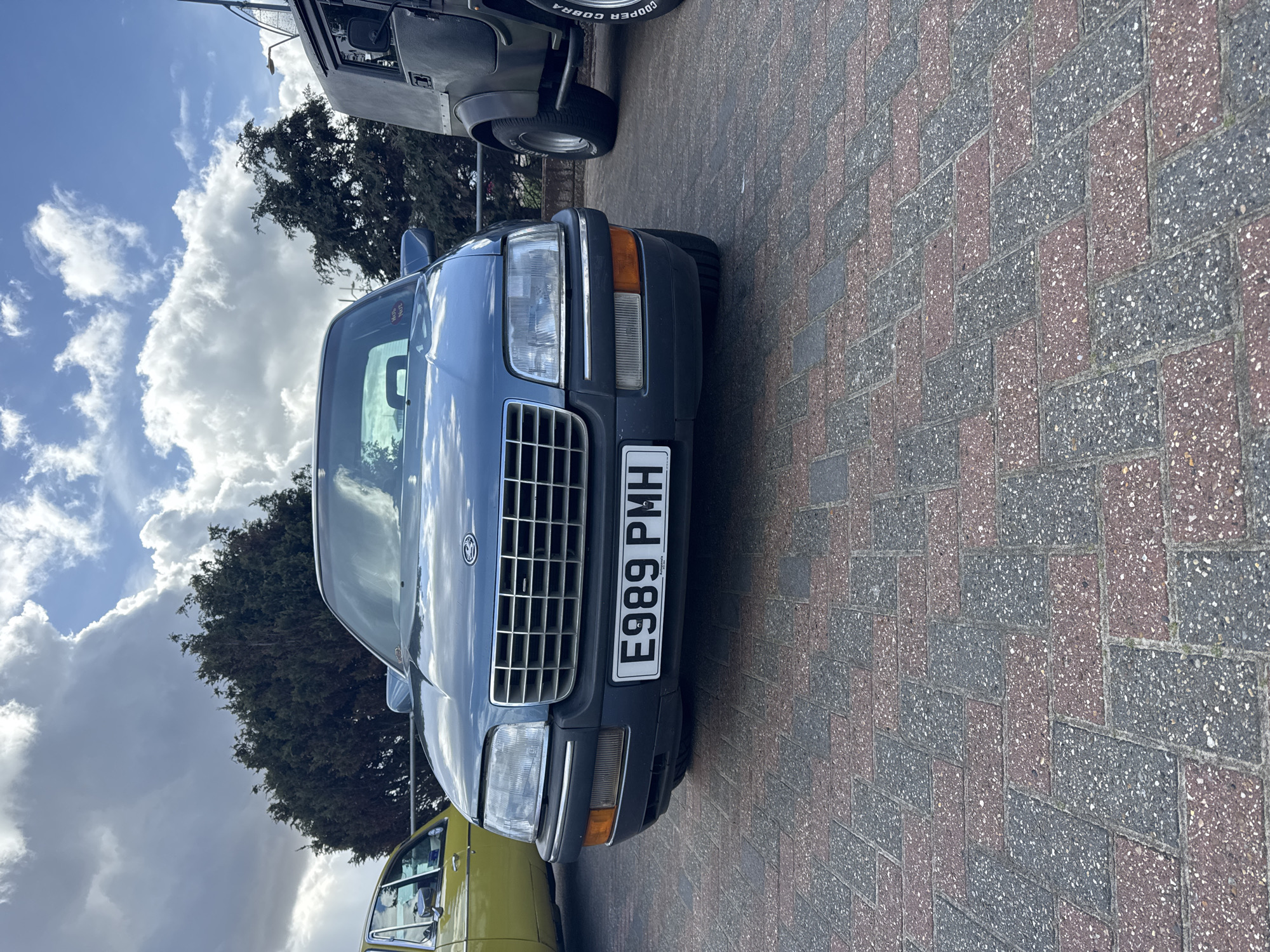 The image shows the front of a dark blue Vauxhall Carlton car, registration number E989 PMH, parked on a paved area with red and grey bricks. The car is facing the camera. A section of a dark gray vehicle, possibly a Land Rover, is visible at the top of the frame. The side of a yellow classic car is also partially visible at the bottom left of the frame.
A tree and a cloudy sky are visible in the background.
The image was taken in Great Yarmouth, United Kingdom.