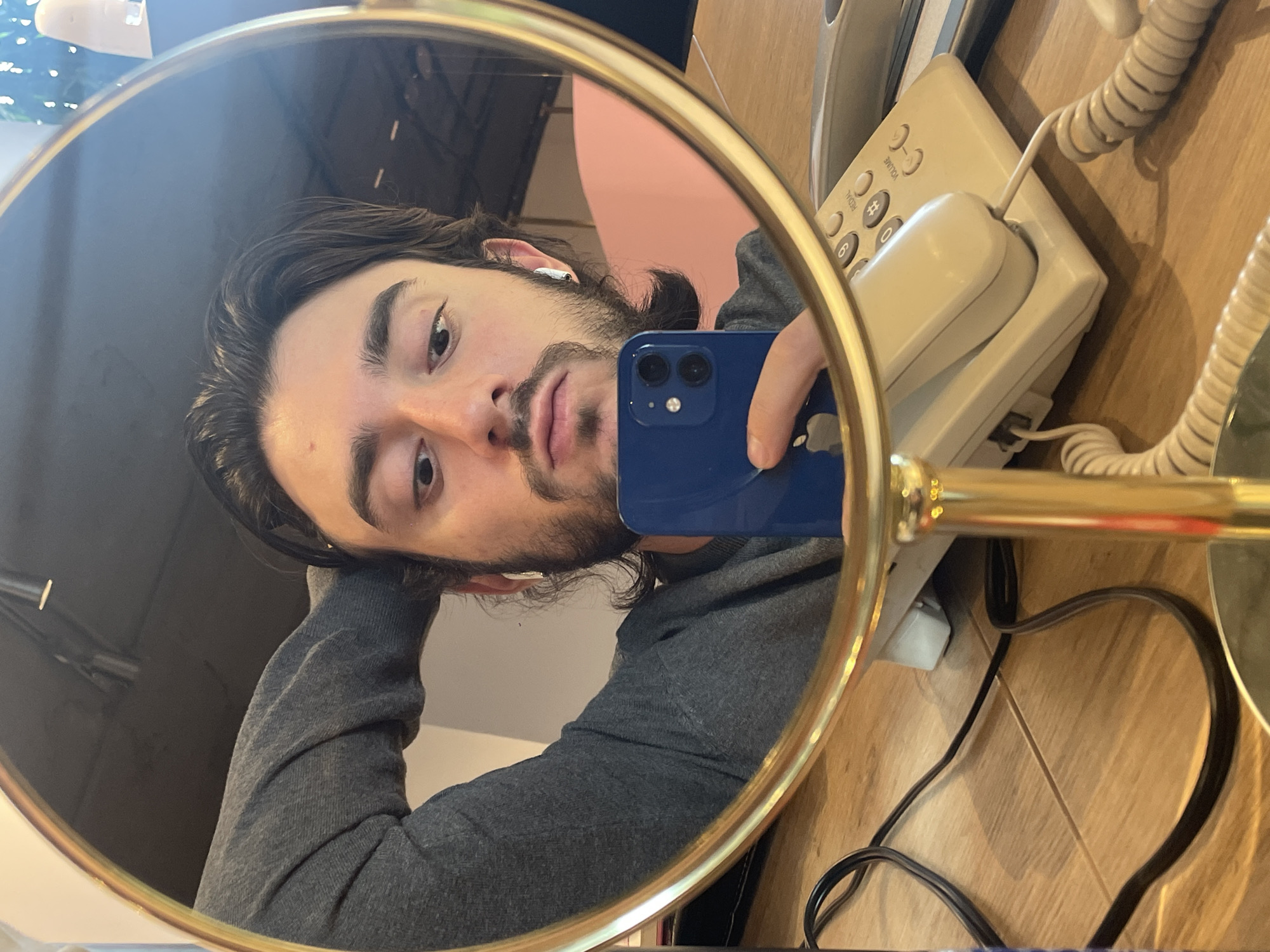 A selfie of a person with dark hair and a beard, wearing a gray sweater and what appears to be a white earbud in their left ear, is captured in a round mirror. The person is holding a blue smartphone with a dual-camera setup. Adjacent to the mirror, a beige telephone sits on a wooden surface. Black electrical cords run along the surface. The selfie appears to be taken indoors. There is no definitive information in the image that confirms the location as Mexico City, Mexico.