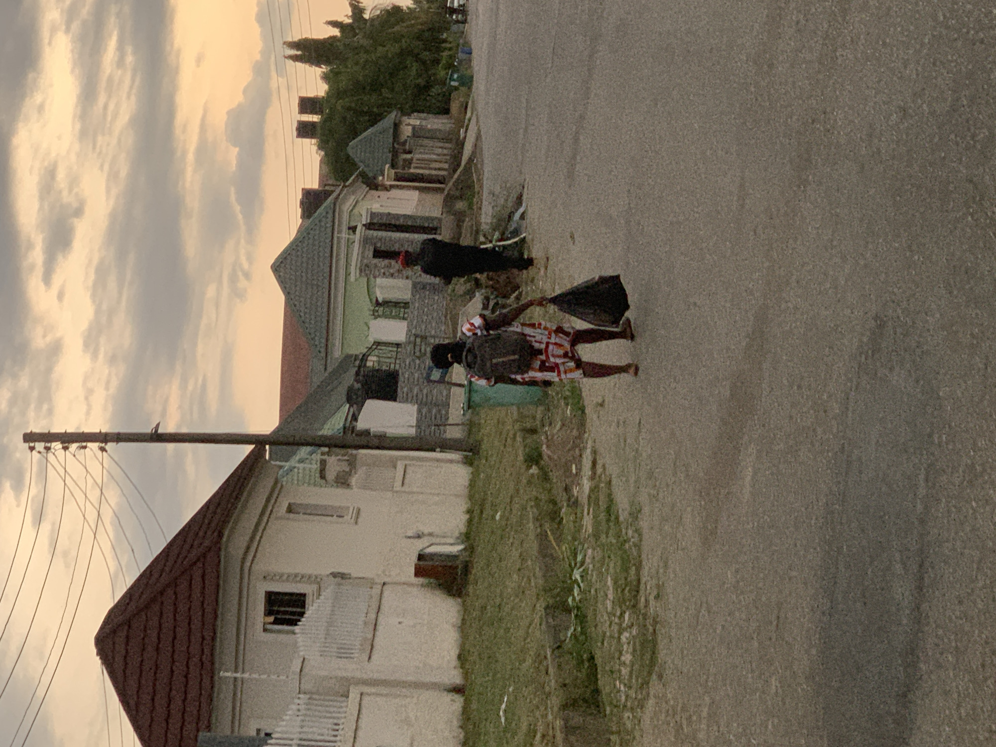 The image depicts a street scene in Abuja, Nigeria, at sunset. The sky is filled with orange-tinged clouds. The street runs diagonally through the frame. Two individuals are visible. One is wearing a black outfit, including a head covering, and is engaged in cleaning the side of the street, while the other is dressed in a colorful patterned outfit and carrying a black bag. Houses with various architectural styles are situated on either side of the street. Utility poles and electrical wires are prominent near the house with the brown roof on the left side of the frame. The street is paved, with patches of grass and debris scattered along the edges.