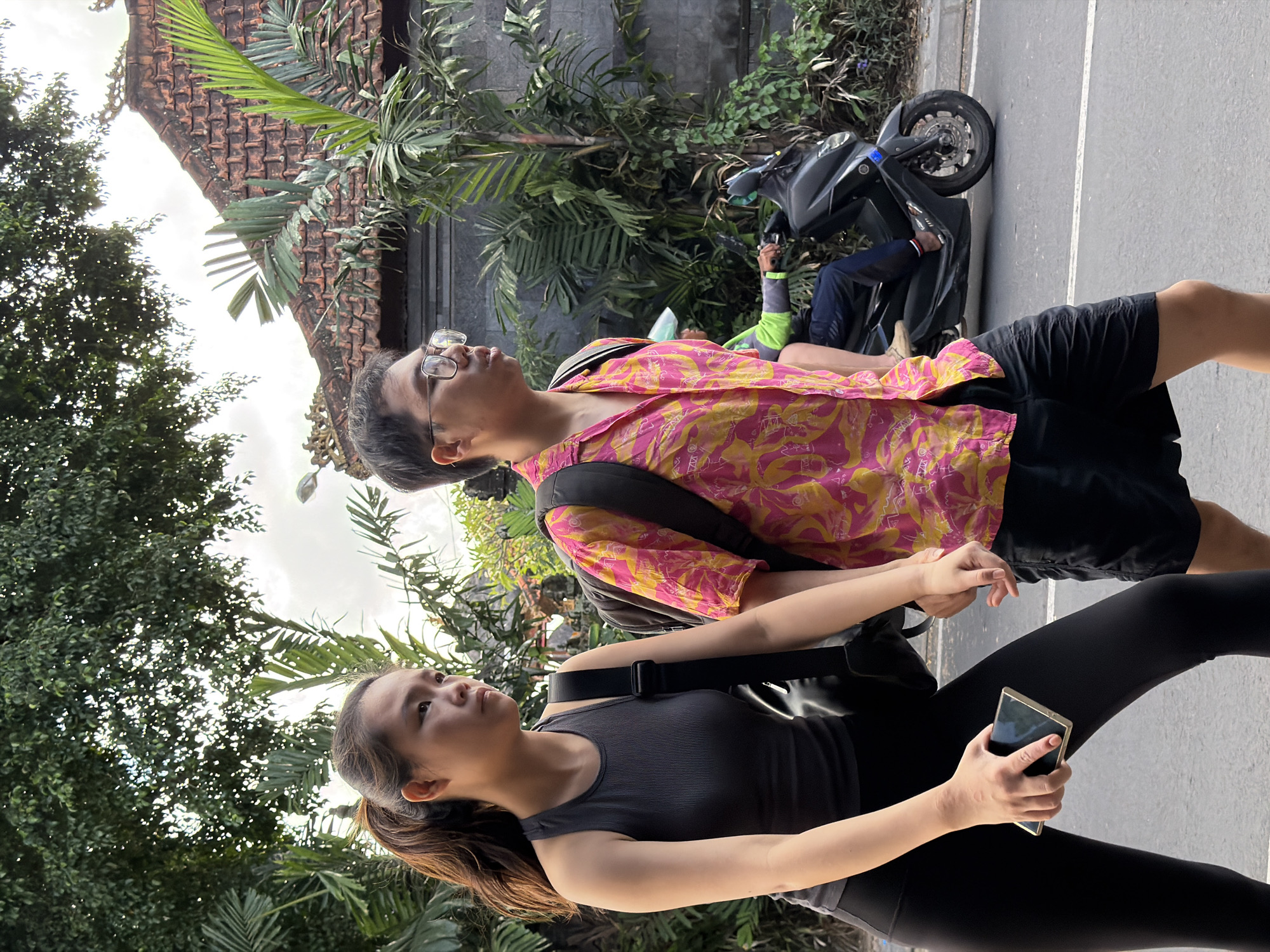 Two individuals, a woman and a man, are walking side-by-side on a paved road. The woman, on the left, wears a dark grey sleeveless top and black leggings. She holds a smartphone in her right hand. The man, on the right, is wearing a bright pink and yellow floral print short-sleeved shirt and black shorts. Both individuals have backpacks. In the background, a person is riding a black scooter. The environment appears to be a street in North Kuta, Indonesia, with lush green foliage, palm trees, and a building with a tiled roof visible behind the pedestrians and scooter. The sky is overcast.