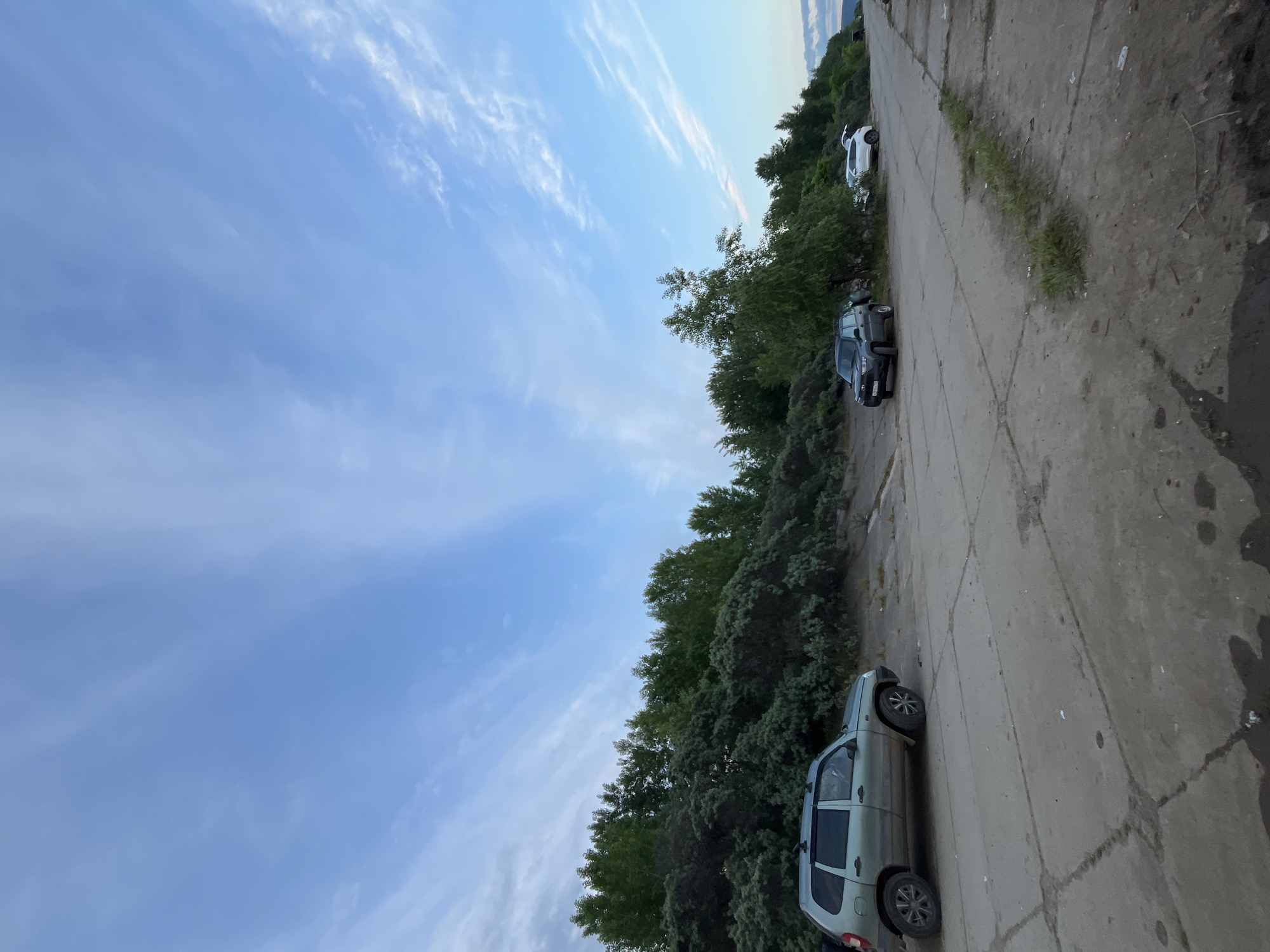The image, viewed upright, captures an outdoor scene featuring a paved road or parking area flanked by dense green foliage on the left and a wide expanse of sky on the right. Several cars are parked along the edge of the paved surface. In the foreground, a light-colored SUV, possibly light green or grey-green, is visible. Further along, a dark-colored SUV is parked, and a white SUV can be seen in the background.
The paved area appears to be concrete or asphalt, showing visible cracks, seams, and some sparse patches of grass growing through. The sky is clear blue with delicate, wispy white clouds stretching across, suggesting fair weather. The light quality indicates late afternoon or early evening. No people are visible, and the scene is static, depicting only parked vehicles. There is no visible text. The setting is consistent with a quiet, possibly semi-rural area in Chaikovsky, Russia.