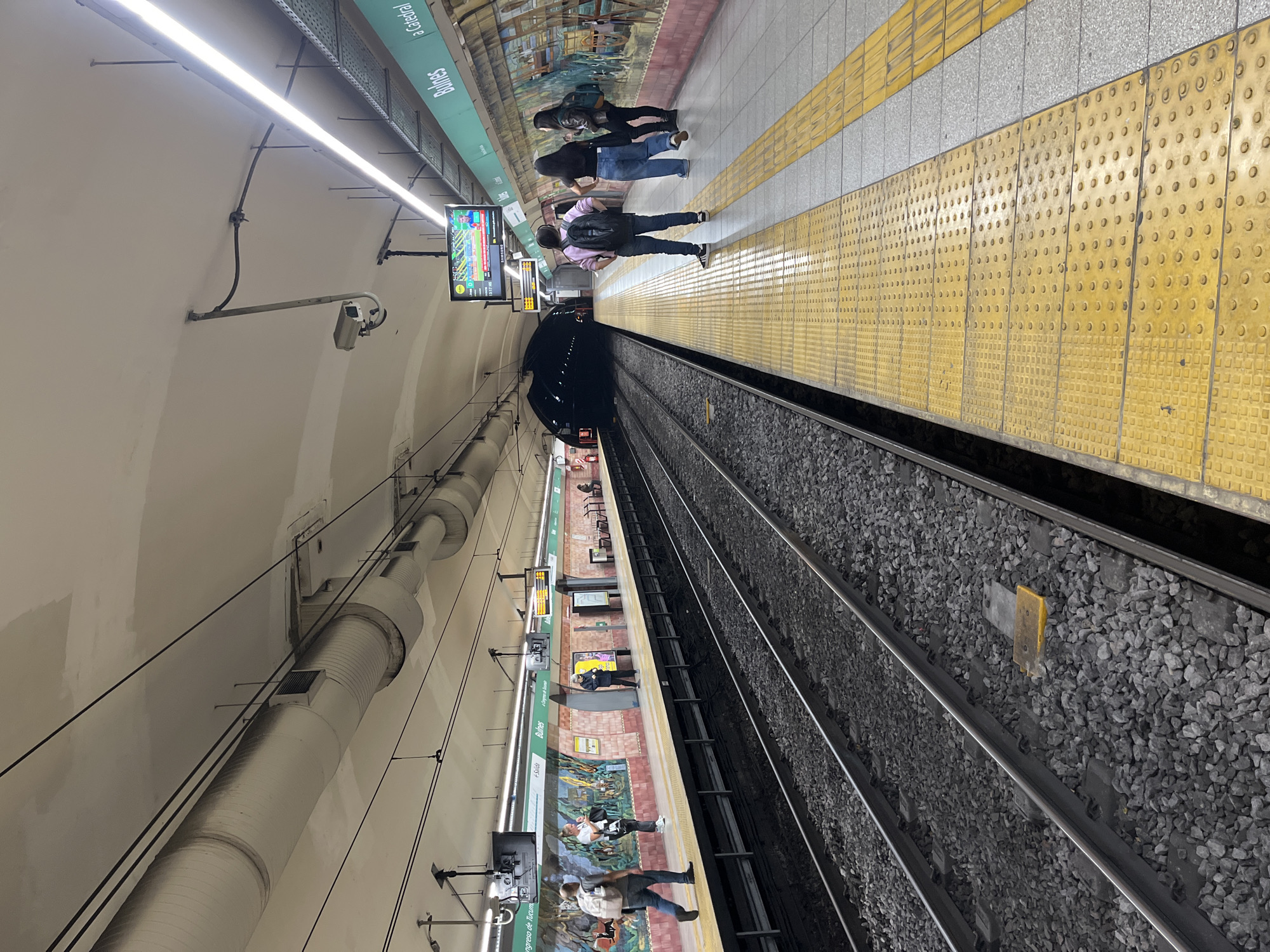 This image captures a vibrant underground subway station platform in Buenos Aires, Argentina, seen from a perspective looking along its length towards a tunnel entrance. The station is brightly illuminated by numerous overhead fluorescent tube lights, casting a warm glow on the arched ceiling and platform.
On the right side, a platform is visible with a prominent yellow tactile paving strip running along its edge, next to the tracks. Several individuals are waiting on this platform. In the foreground, a person wearing a pink shirt and a black backpack stands facing the tracks, seemingly awaiting a train. Behind them, two other people are also standing and looking towards the tracks. Further down the platform, more passengers are spread out, some standing and others possibly seated on benches on the opposite platform, which is partially visible across the tracks.
The platform wall is adorned with a colorful, detailed mural depicting various scenes, adding an artistic touch to the utilitarian setting. Above the platform, digital display screens show information; one appears to be displaying what looks like sports scores or a broadcast, while another has numerical information, possibly indicating train arrival times. Visible text on signs includes the station name "Buines," and directional signs "a Catedral" and "Congreso de Tucumán," with an arrow pointing to an exit labeled "← Salida."
Two sets of train tracks, filled with grey gravel ballast, stretch away from the foreground and disappear into a dark tunnel opening in the distance. The ceiling features large, silver-colored ventilation ducts and various electrical conduits and security cameras mounted along its curved surface. The atmosphere is calm, with people patiently waiting for their transport.