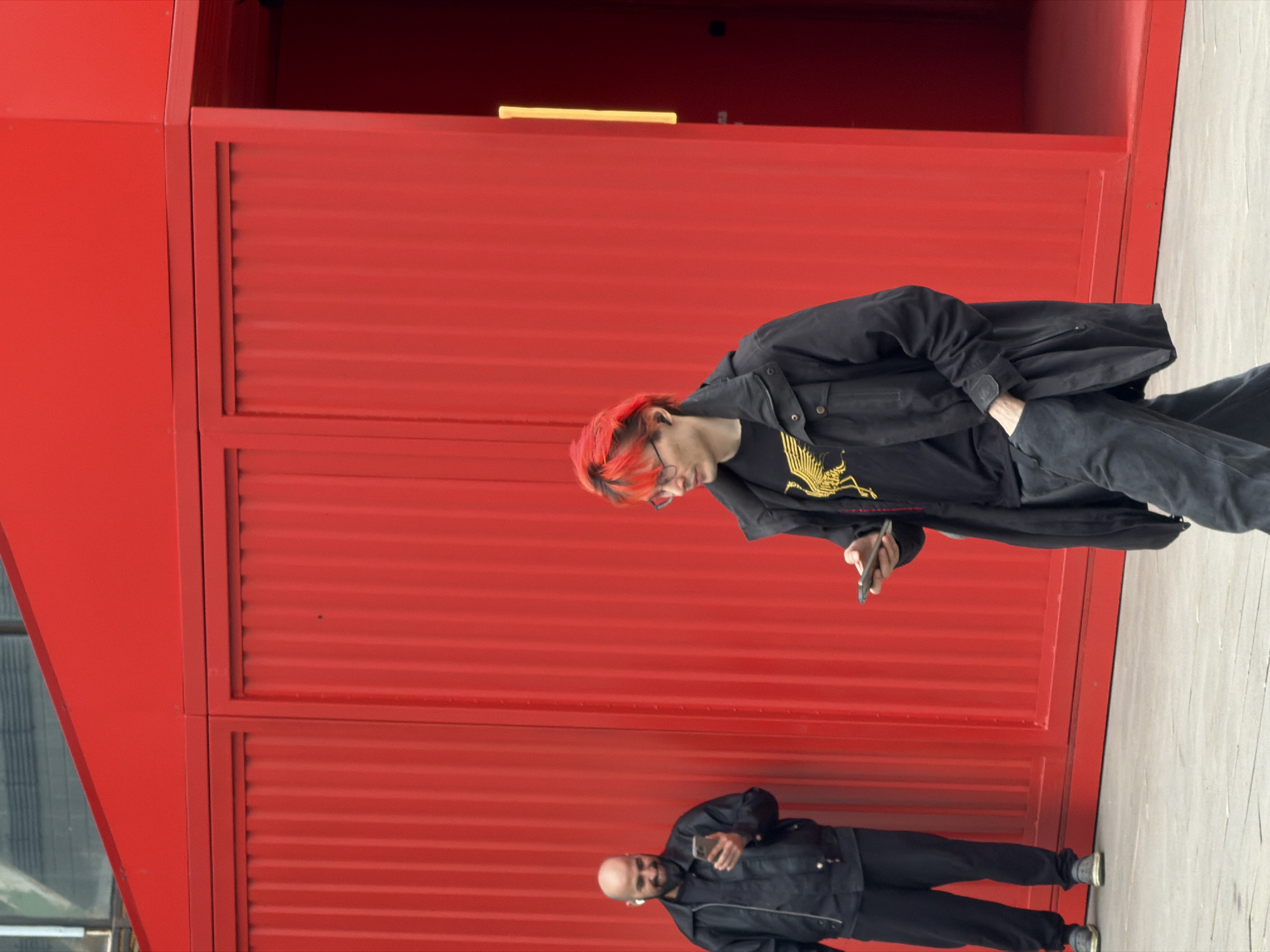 The image shows two people standing in front of a bright red, corrugated metal structure. One person, with red hair and glasses, is looking at their phone. They are wearing a black jacket and a t-shirt with a yellow design. Below them is another person, bald and bearded, also looking at their phone and wearing a black jacket and pants. The ground appears to be concrete.
Given the architectural style and available information, the scene could be located in Saint Petersburg, Russia, near a modern building or art installation.