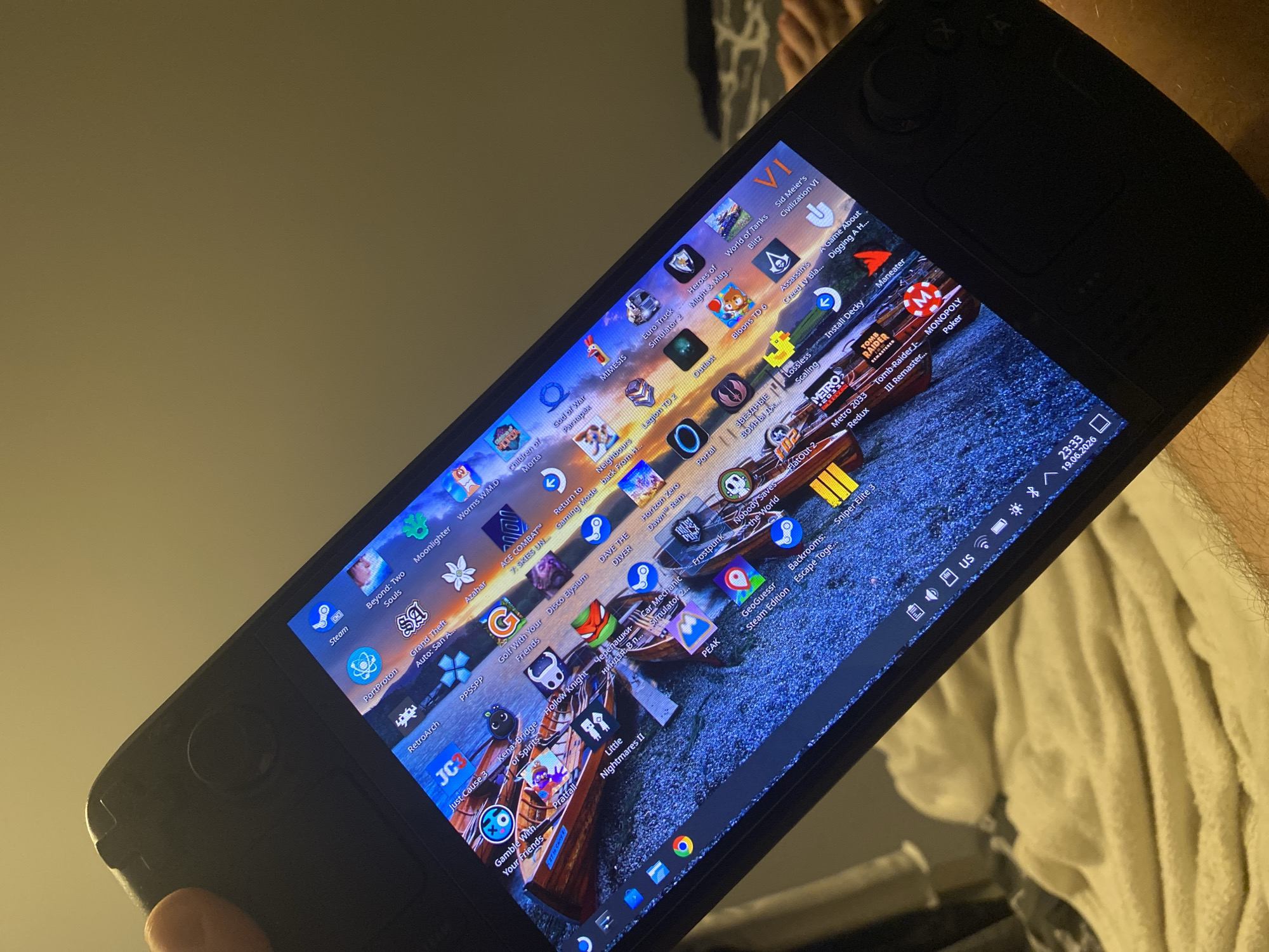 The image shows a close-up, angled view of a person's hand holding a black handheld gaming device, likely a Steam Deck, resting on a white, textured surface. The device's screen displays a cluttered desktop environment with numerous game icons and a portion of the taskbar visible at the bottom. The wallpaper appears to be a scenic image of canoes on a body of water with rocky shores. The background is out of focus, suggesting an indoor setting with warm, ambient lighting. No specific location in Łódź, Poland is discernible from the provided image.