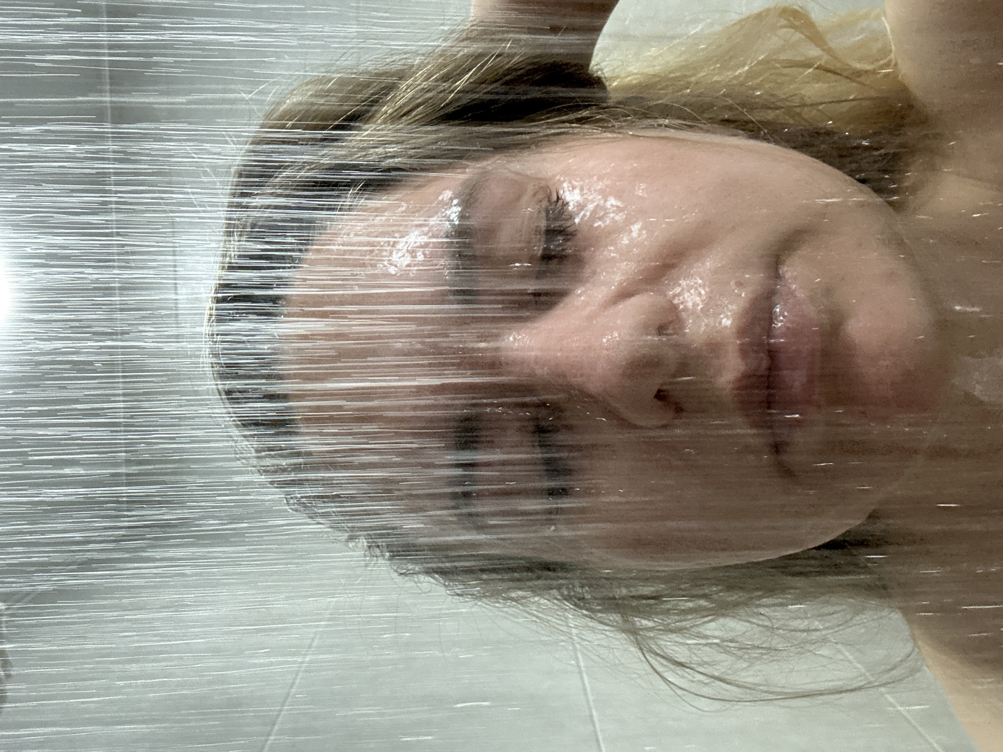 A close-up, indoor shot depicts a fair-skinned person's face obscured by what appears to be translucent wrapping material. The background includes tiled flooring, possibly in a bathroom or similar setting. The perspective is slightly angled, suggesting the photo was taken from a higher vantage point. The location information (Chaikovsky, Russia) is insufficient to provide additional contextual details.