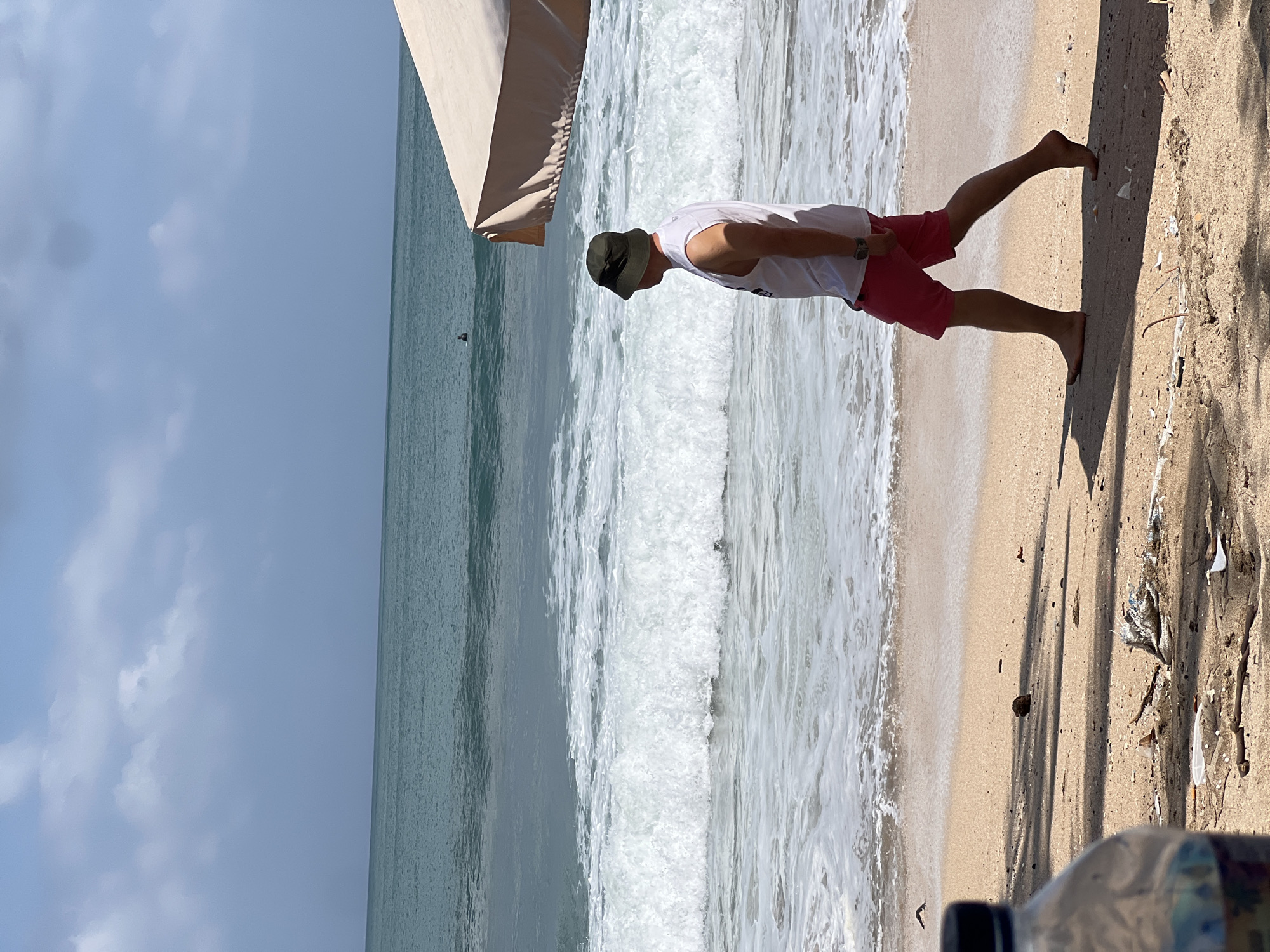 A man wearing a white tank top and red shorts walks along the sandy shore of Kuta Beach in Bali, Indonesia. He is also wearing a dark bucket hat and a watch on his left wrist. The ocean stretches out behind him, with waves crashing onto the beach. The sky is partly cloudy. To the right of the frame, part of a large, light-colored umbrella is visible, casting a shadow on the sand. The foreground shows a portion of a plastic water bottle and scattered debris on the beach, including plastic waste and dried plant matter. A lone surfer is visible in the distance, on the ocean.