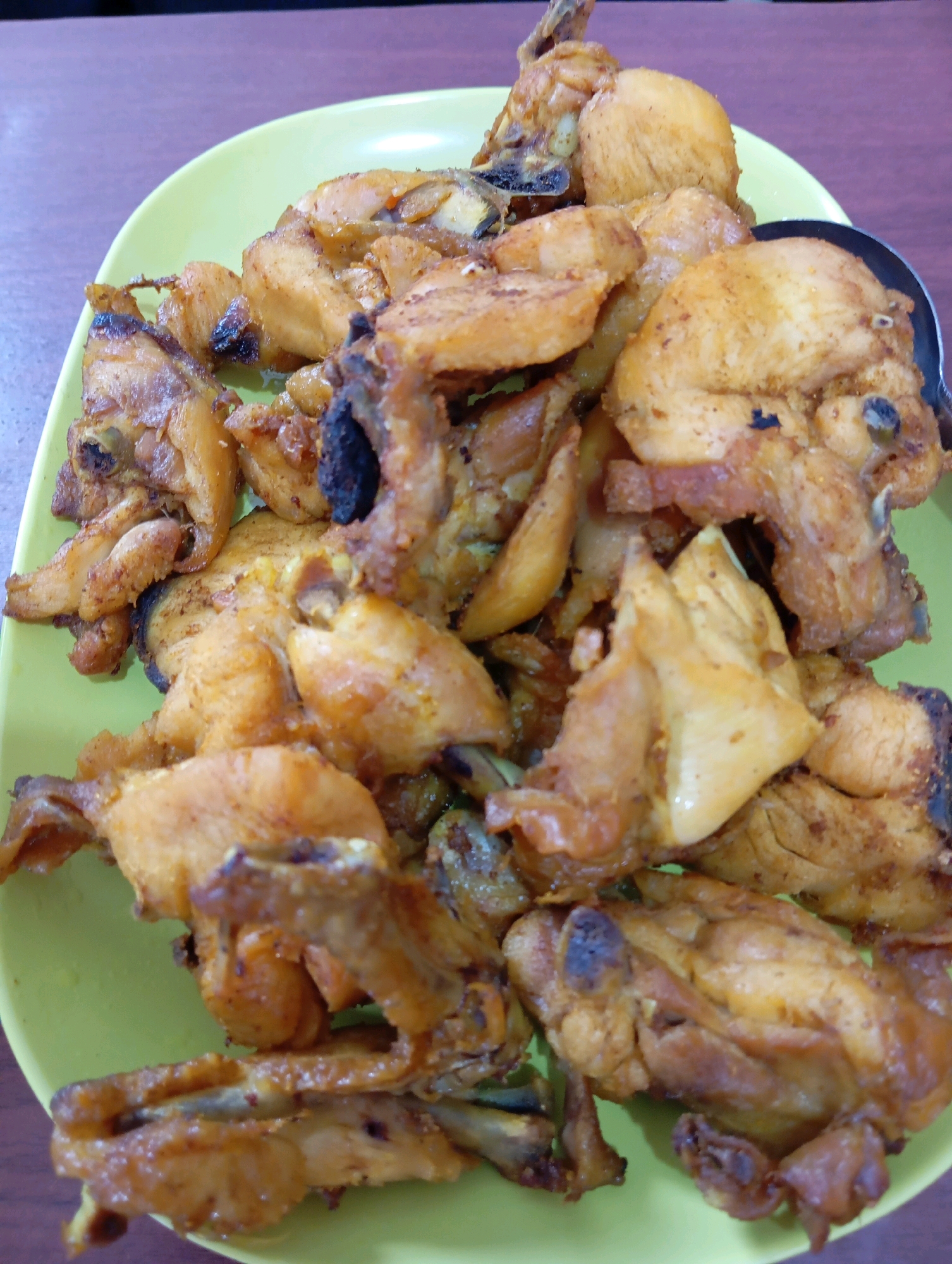 A close-up, overhead shot features a bright green plate filled with pieces of fried chicken, positioned on a brown wooden table. The chicken pieces are varied in size and shape, some appearing crispy and others more tender. They are arranged in a pile, obscuring most of the plate. A silver spoon is partially visible on the right side of the plate, resting on top of some of the chicken. The lighting is consistent across the image, casting soft shadows. The background is out of focus, providing context but not distinct detail. The scene appears to be a casual dining setting. The city of Curah Kalong, Indonesia is not identifiable from the visual information provided.