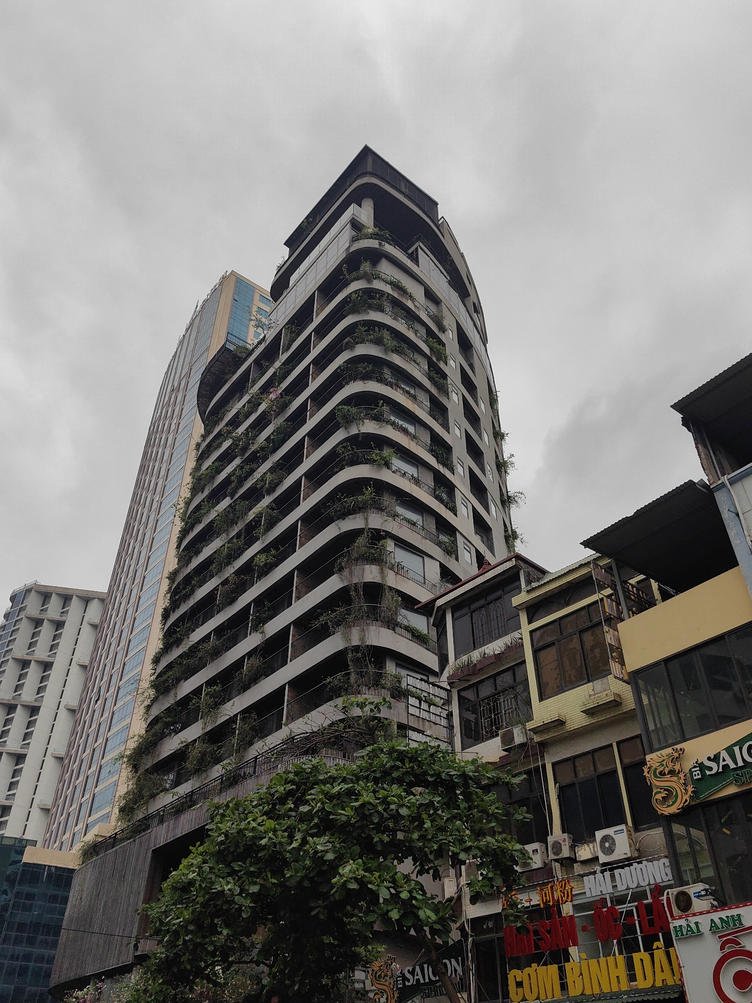 The image shows a street scene in Bai Chay Ward, Vietnam. A tall, curved building with vegetation growing on its balconies dominates the frame, standing alongside other high-rise buildings. In the foreground, there are smaller buildings with signs in Vietnamese, including a sign for "SAIGON." Air conditioning units are visible on the facades of these buildings. A tree is present in the lower center of the image. The sky is overcast, suggesting cloudy weather.