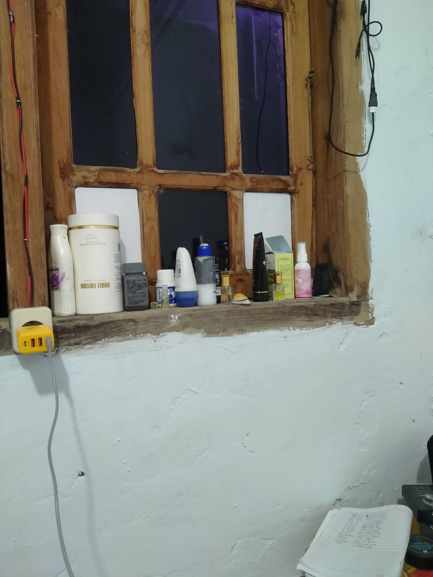 The image depicts a cluttered windowsill in a room, likely in Blaban, Indonesia. The windowsill itself is made of weathered wood and is covered with various personal care items, including bottles, jars, and tubes. Among the items are a large white container with "NOBLE EDITION" printed on it, a smaller white bottle with a floral design, a grey bottle with Chinese text, a white spray bottle, and other miscellaneous containers.
To the left of the windowsill, a yellow USB charger with multiple ports is plugged into a white wall socket. A gray cable extends from the charger and dangles down. On the far left, a black and red electrical wire runs vertically along the wooden window frame.
The window behind the windowsill is divided into multiple panes by wooden mullions. The glass panes are dark, suggesting either tinted glass or that it is nighttime. The wall to the right of the window is painted white and shows signs of wear, with peeling paint and textured imperfections.
In the lower right corner of the image, a notebook with handwritten text is visible, along with a portion of a black object that could be a calculator or another electronic device. A small dark green container is also partially visible. The overall impression is one of a lived-in space with a functional but unorganized arrangement of everyday items.