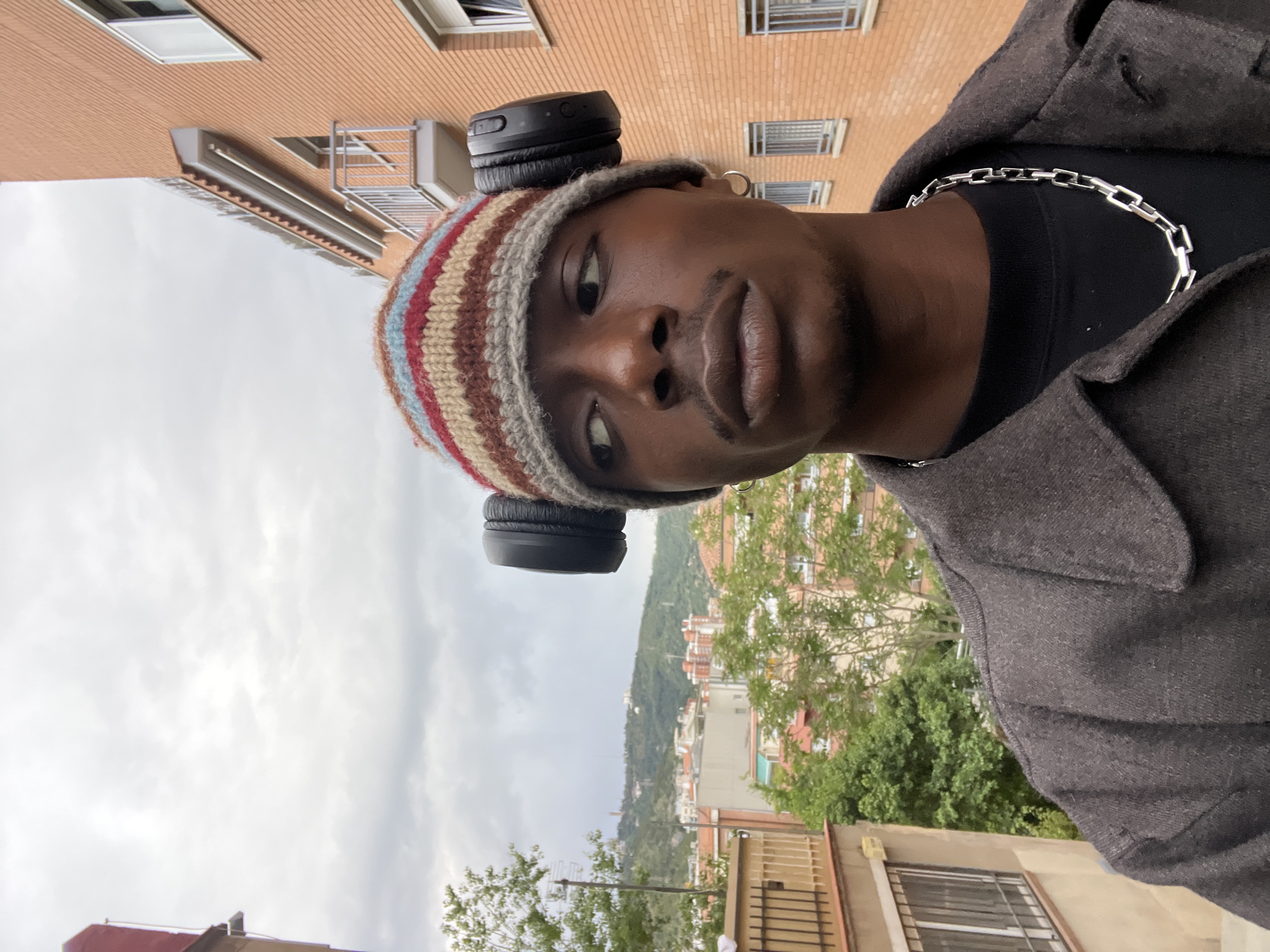 A medium-angle, close-up, portrait shot shows a Black male wearing a striped beanie with black headphones on top of it. The man is also wearing a gray coat and a silver chain around his neck. The location appears to be Barcelona, Spain, given the architectural style of the buildings in the background and the presence of green hills in the distance. The sky is cloudy and gray. The image is rotated counterclockwise.