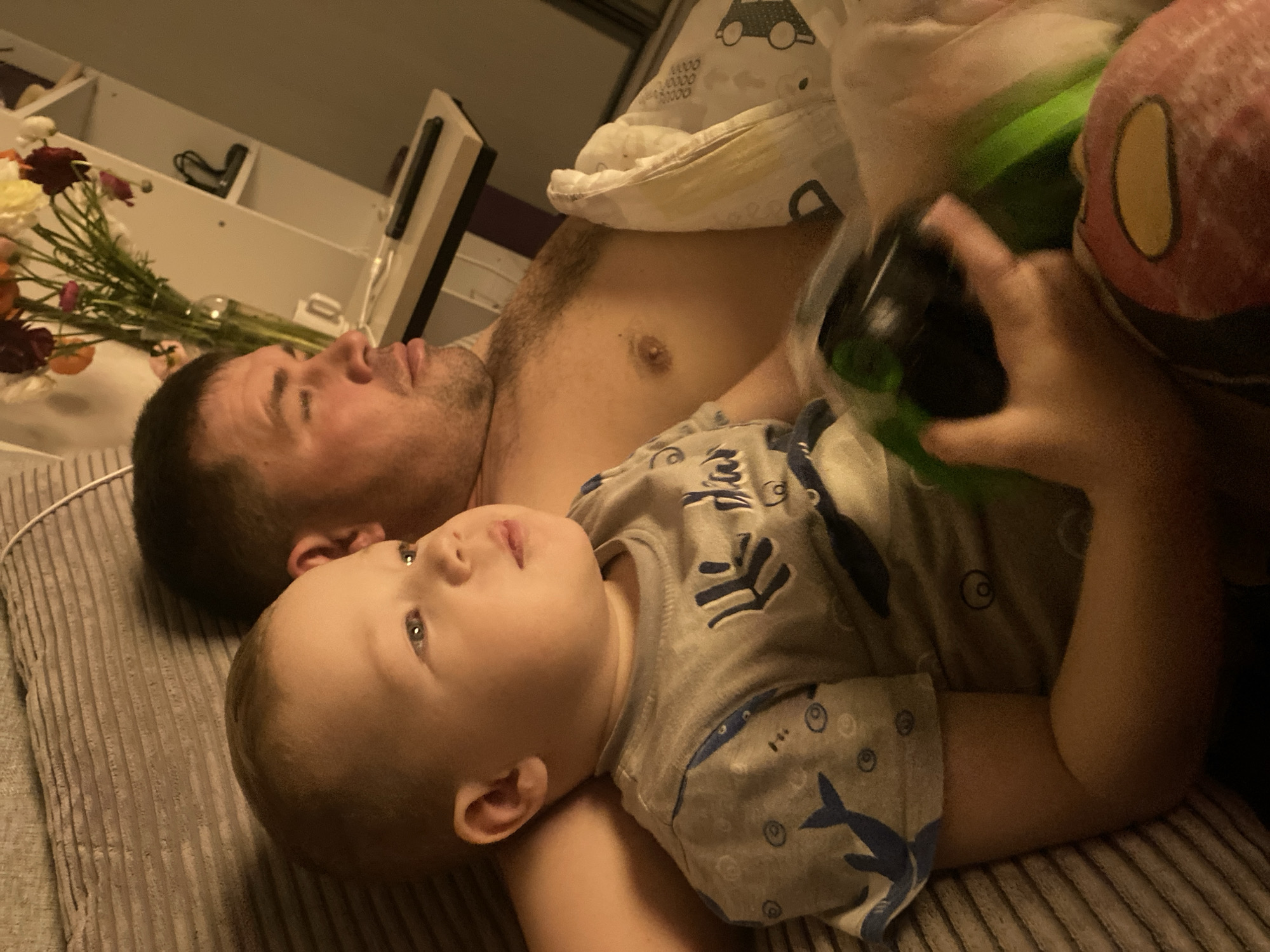 This image captures a serene, intimate moment between an adult male and a young child, likely a father and son, resting together indoors. The scene appears to be set in a bedroom, characterized by the bedding and relaxed postures.
The man, positioned on the left side of the frame, is shirtless and appears to be asleep or deeply resting, with his eyes closed or nearly closed. He has short, dark hair and light stubble on his face, conveying a sense of calm. A white blanket adorned with car patterns is partially draped over his chest.
Lying closely beside the man, towards the bottom right, is a young child, likely a toddler. The child is awake and looks upwards and slightly to the left with an alert and curious expression. They are wearing a light-colored, long-sleeved top patterned with blue sharks and circles. The child's right hand holds a dark-colored object, possibly a bottle or a small toy. A pink and yellow plush toy is also partially visible on the far right.
Both individuals are resting on a ribbed, light brown or beige textile, possibly a duvet cover or blanket. In the background, to the left, a vase containing an assortment of flowers (red, white, and orange) is visible on a surface. Above this, a light-colored shelf unit with some indistinct items can be seen. The lighting in the image is dim, suggesting it might be late evening or early morning, contributing to the quiet and peaceful ambiance. There is no visible text in the image. The location context provided for this media is Sochi, Russia.