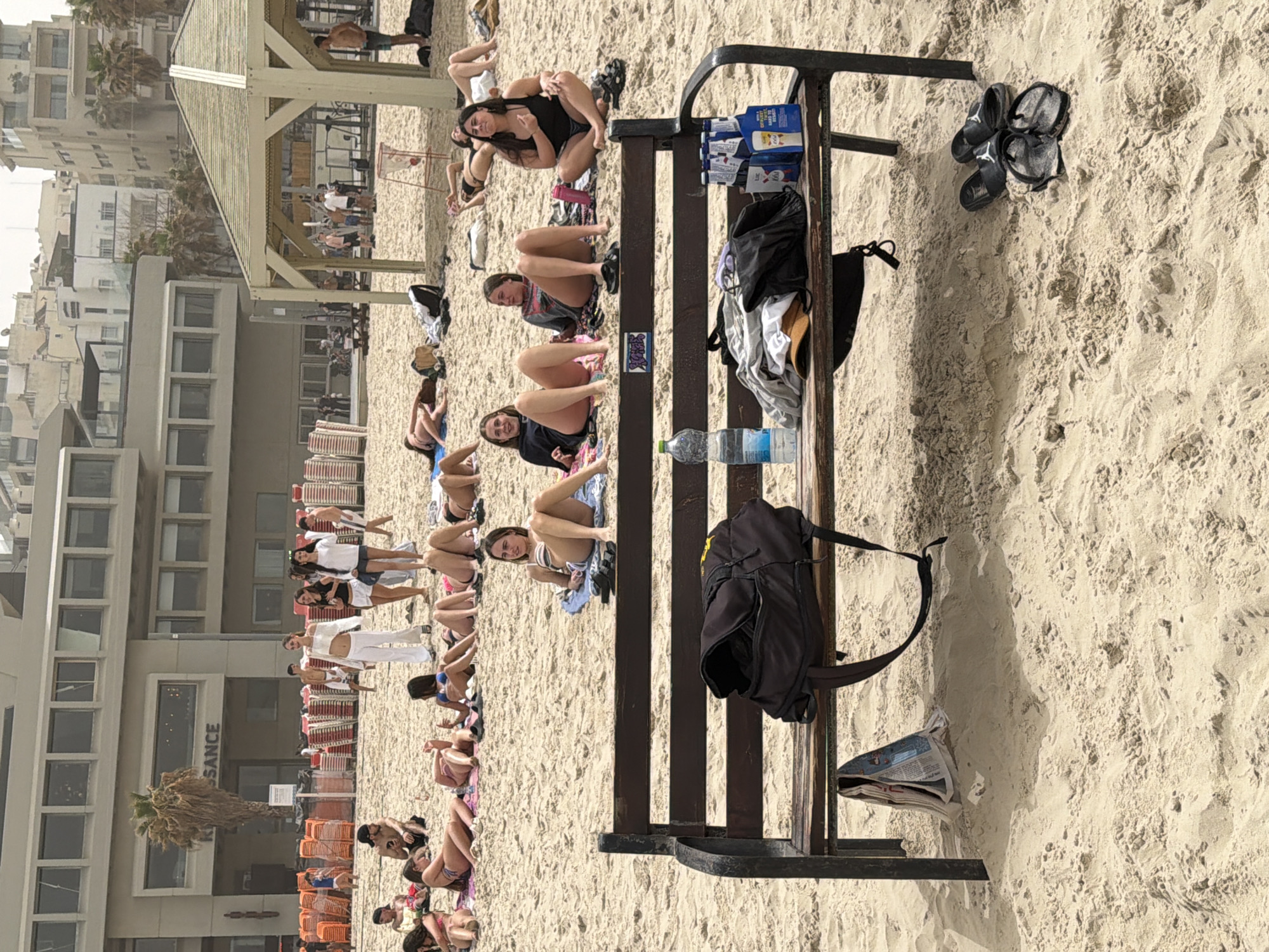 The image captures a beach scene in Tel-Aviv, Israel, with a row of people sunbathing on the sand near a building labeled "RENAISSANCE". A wooden and metal bench stands prominently in the foreground, holding a backpack, a water bottle, a newspaper, packs of liquid, and clothes. Two pairs of flip-flops rest on the sand next to the bench. In the background, more people are lying on the beach under a shaded structure.