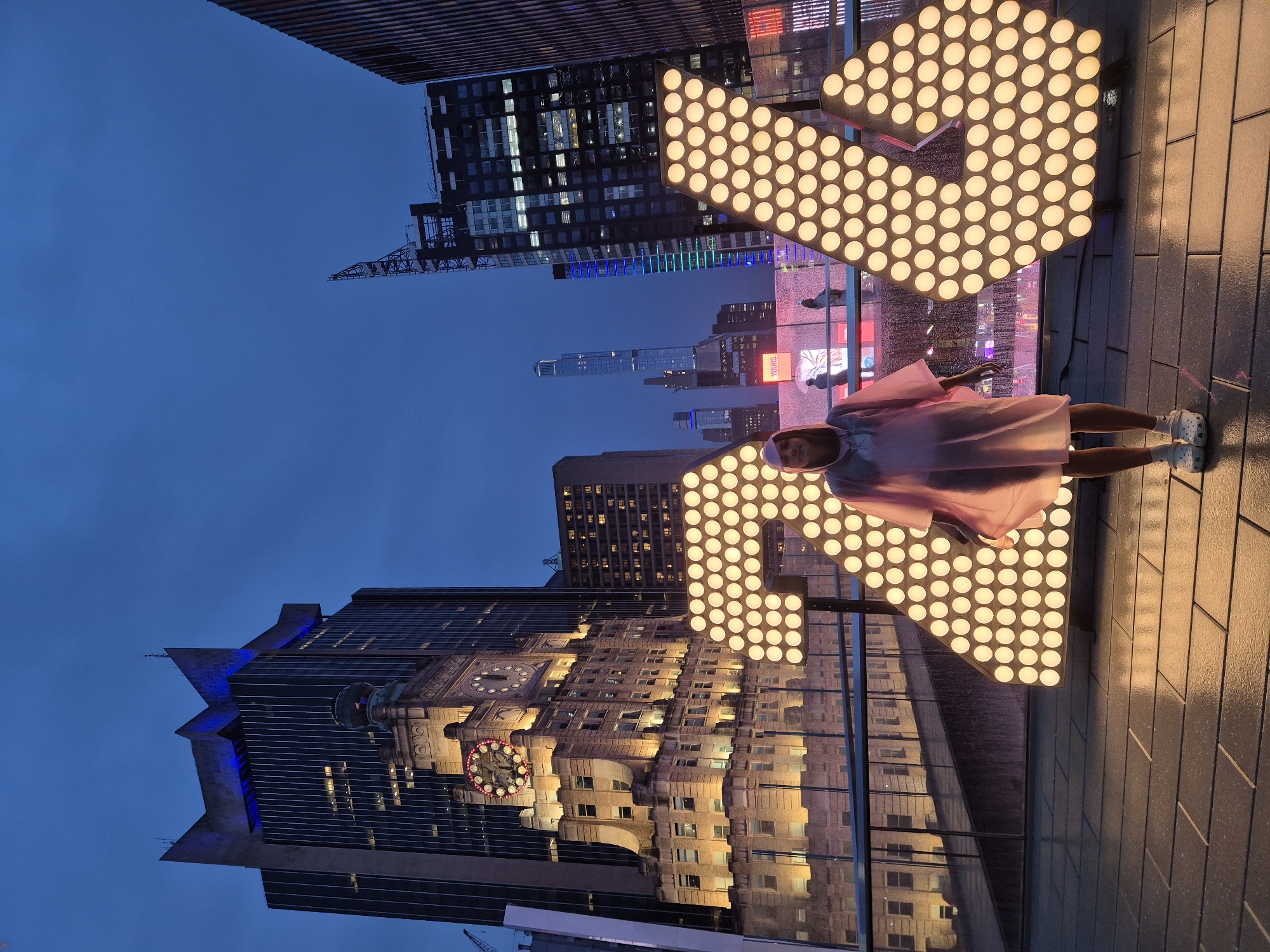 A person stands in front of a large, illuminated number "24" installation in Times Square, New York City, USA, at dusk. The person is wearing a pink poncho and white Crocs. The background features several tall buildings, including the iconic Times Building with its illuminated clock face. The sky is a deep blue, indicating it is twilight. The "24" installation is made of numerous bright, circular lights. The ground is paved with dark tiles. The scene suggests a nighttime event or celebration in a bustling urban environment.