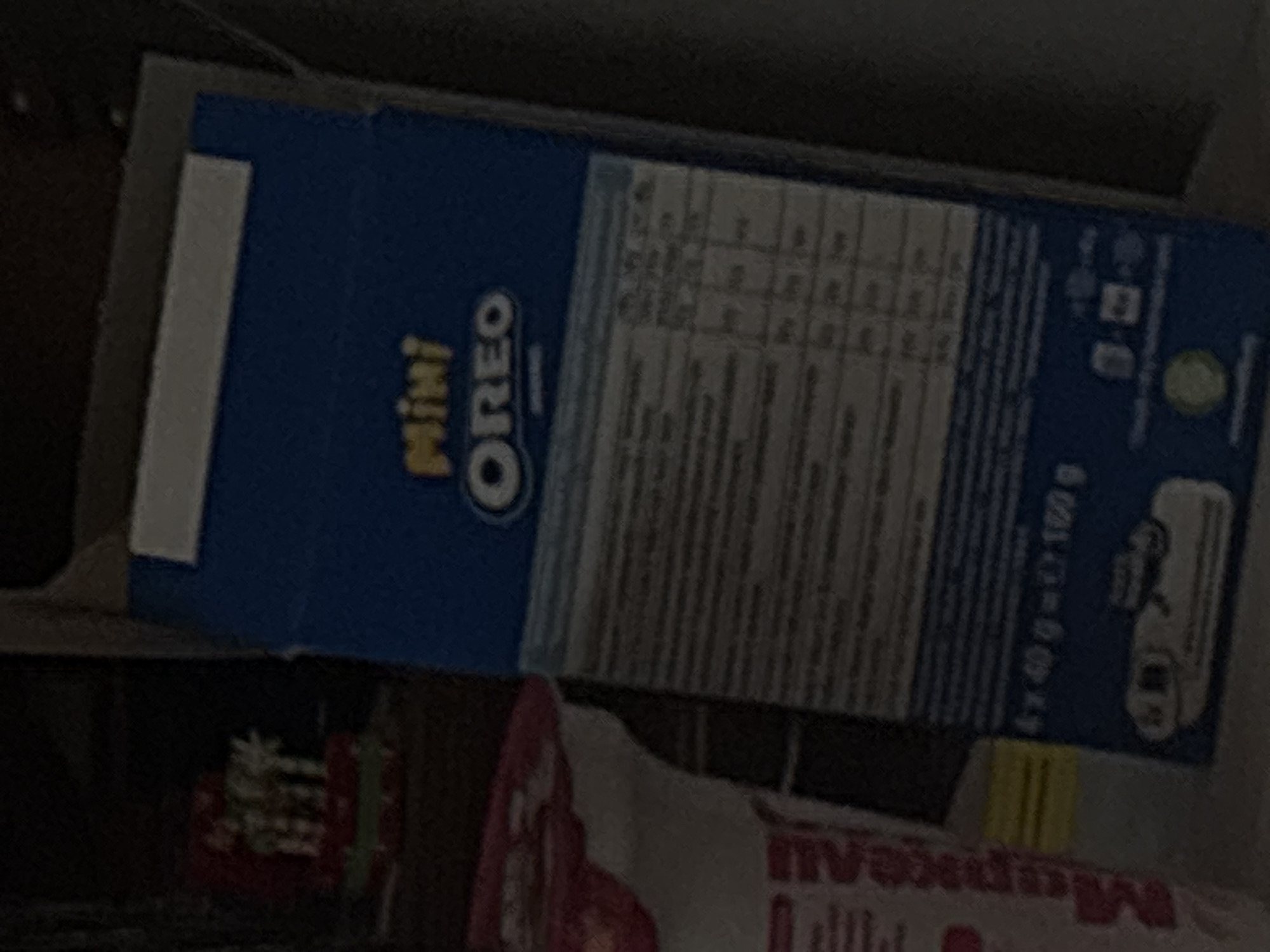 The image displays an indoor scene characterized by low light conditions, situated within the city of Burgas, Bulgaria. The central object is a vertically oriented blue cardboard box, identified as "Mini OREO" by prominent yellow and white lettering near the top. Below the brand name, a grid-like table containing indistinct text, likely nutritional information, is present but largely unreadable due to image blur and underexposure. Near the bottom, the packaging explicitly states "4 x 40 g = 160 g", indicating the total weight and number of individual portions. Several small, unidentifiable logos or symbols, including a discernible green circular recycling emblem, are located at the bottom edge of the box.
To the left of the blue box, a white container, possibly a cup or tub, is partially visible. This container features vertical red or pink text, with partial Cyrillic characters that appear to spell "Мержеви" along its side. Further left in the background, an out-of-focus decorative item resembles a miniature festive cake or ornament, displaying white, candle-like elements atop a base with red and green accents. The overall environment is dimly lit and appears to be a domestic setting. No individuals are present in the image, and no actions or interactions are depicted. The precise location within Burgas, Bulgaria, beyond the general city context, cannot be determined from the visual information provided.