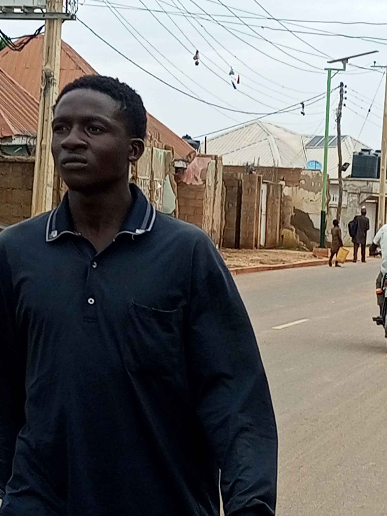 In a street scene in Tempure, Nigeria, a young Black male, appearing to be in his late teens or early twenties, walks towards the left of the frame. He is wearing a dark blue long-sleeved polo shirt with white and grey trim around the collar and a single white button visible at the neckline, as well as a breast pocket. His dark hair is short and styled. Behind him, the street is paved with asphalt and marked by a white line. To the left of the street, buildings with corrugated metal roofs are visible, some appearing weathered. Utility poles with numerous electrical wires stretch across the sky. Further down the street, a small child, also Black, walks away from the camera, carrying a yellow container. Another person, a Black male dressed in dark clothing and a head covering, stands near the child. The sky is overcast. The overall impression is of a typical urban environment in Nigeria.