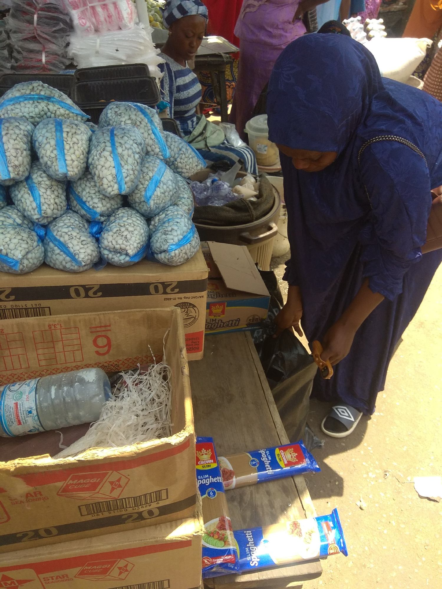 This media file shows a busy marketplace scene in Jalingo, Nigeria. The primary subjects are two women, likely vendors, interacting with the goods they are selling. One woman, wearing a blue hijab and a patterned blue dress, is focused on something out of frame, possibly bagging or sorting items, and is holding a small orange tool, possibly a knife or scissors. The other woman, partially visible in the background, wears a blue and white striped garment and a headscarf.
The scene is filled with various objects for sale. Prominently displayed are large bags of white beans tied with blue string. In the foreground, there are cardboard boxes containing packaged goods. One box is labeled with "MAGGI CUBE STAR SEASONING" and "20". Another box, partially visible, also has "MAGGI CUBE STAR SEASONING" and "20". There are also packages of "CROWN SLIM Spaghetti". A plastic water bottle is visible in one of the boxes, along with shredded paper.
The setting appears to be an outdoor market, with goods displayed on tables and on the ground. The lighting suggests it is daytime, likely a sunny day, as indicated by the strong shadows. The overall atmosphere seems industrious, with the vendors actively engaged in their work. The presence of staple food items like beans and spaghetti suggests a focus on everyday consumables.