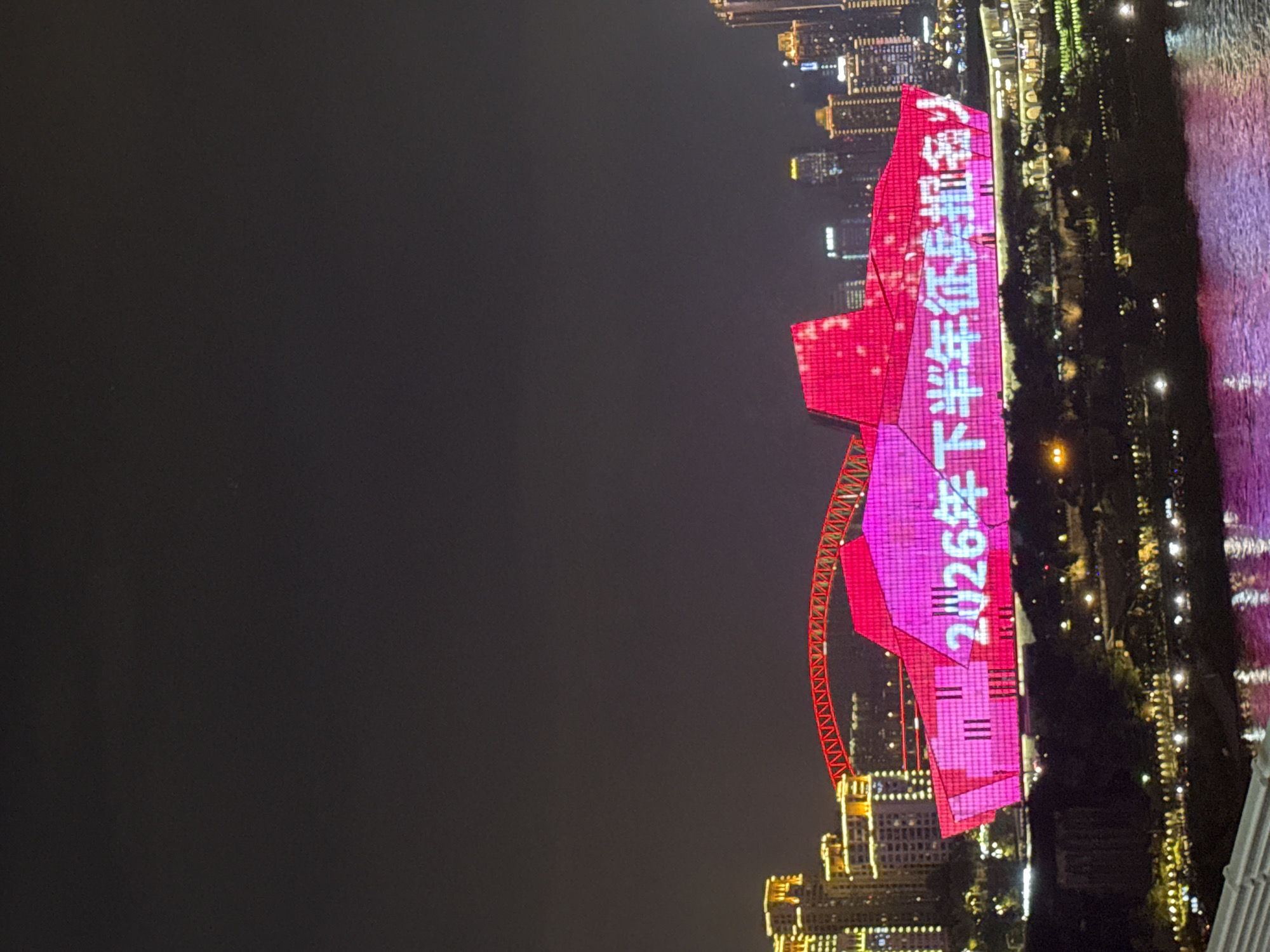 The image captures a nighttime cityscape in Yuzhong District, China, dominated by a large, brightly lit building that appears to be illuminated with a pink LED display. The display shows white Chinese characters that read "2026年下半年征兵报名," which translates to "2026 Second Half Recruitment Registration."
The building itself has a unique, angular architectural design, resembling a multifaceted structure or perhaps a stylized representation of a ship. A red, illuminated archway or bridge-like structure extends from the left side of the building.
In the background, other city buildings are visible, some lit with yellow and white lights, contributing to the overall urban night scene. The lower portion of the image shows a body of water, likely a river, reflecting the city lights and the pink illumination from the main building. The reflections on the water appear as streaks of pink and purple light. The sky is dark, typical of a nighttime photograph. There are no discernible people in the image, but the presence of the lit-up buildings and the river suggests an urban environment with potential public spaces.