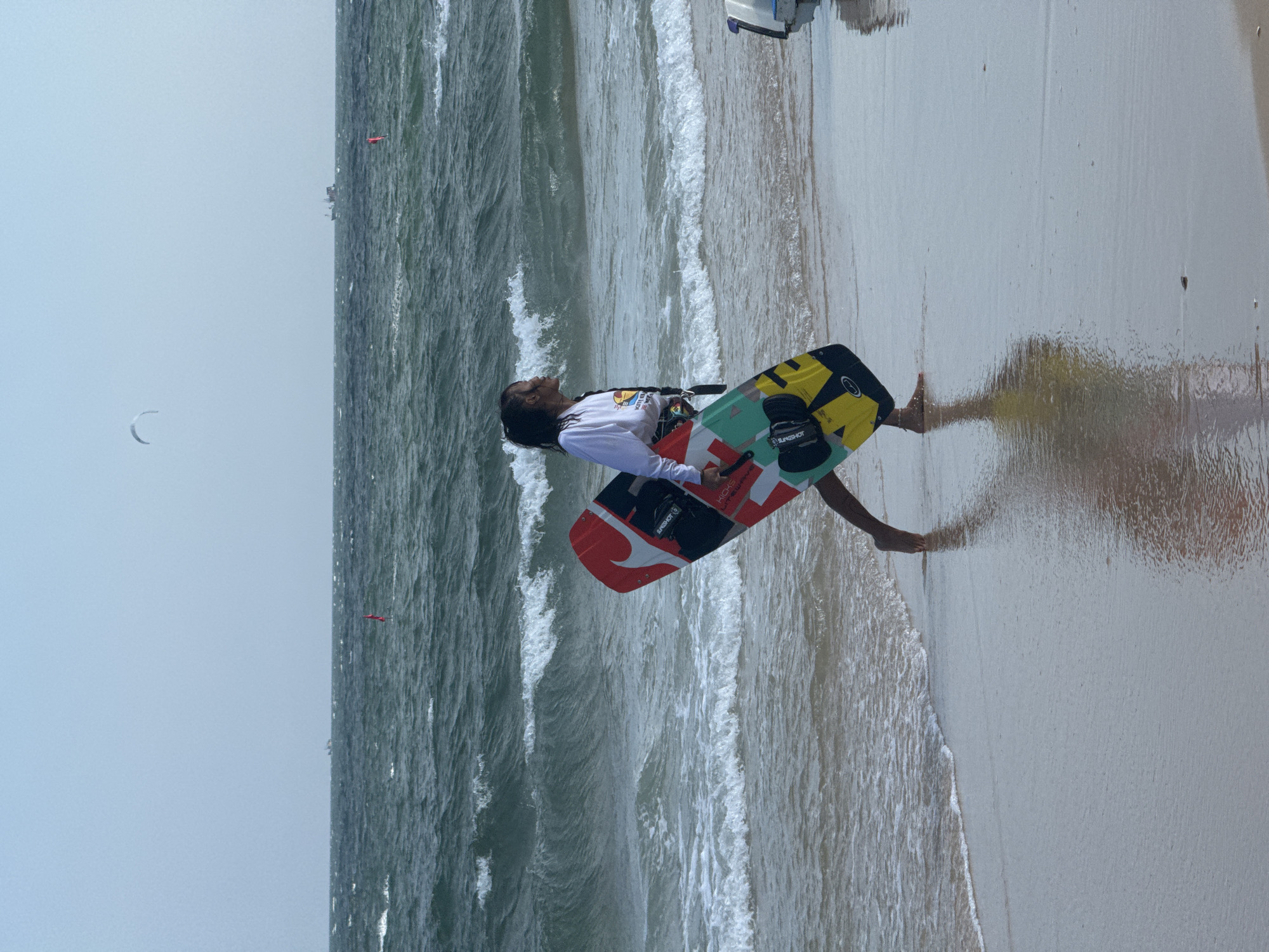 A person walks along the shore of a beach in Phan Thiết, Vietnam, carrying a colorful kiteboard. The sky is overcast, and the sea is choppy with moderate waves. In the distance, a few boats are visible on the horizon, and a kite can be seen in the sky. The person is wearing a long-sleeved shirt and shorts. The kiteboard is red, black, yellow, and green, with the brand "Slingshot" printed on it. The sand is wet and reflective from the receding tide.
