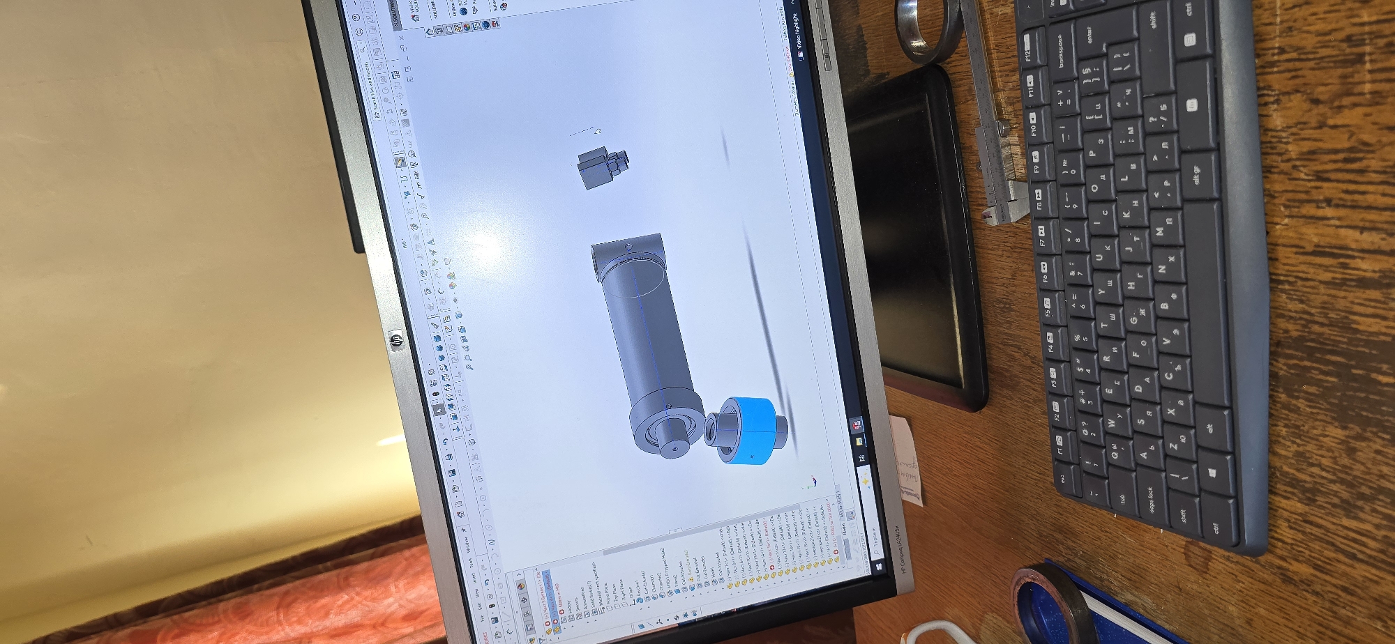 Here's a detailed description of the image:
The image captures a workspace, showing a monitor displaying a 3D CAD model likely created in SolidWorks software. The model appears to be a cylindrical object with attachments, highlighted in blue.
To the right of the monitor, there's a black keyboard. A measuring tool or calipers and a metal ring are also visible near the keyboard.
The environment includes a wooden surface beneath the monitor and keyboard. Part of a reddish/orange blanket is visible to the lower left corner of the screen. A secondary monitor is visible next to the main monitor. A post-it note is stuck to the side of the screen.
There is no information that suggests a location in the city of Komuniga, Bulgaria.