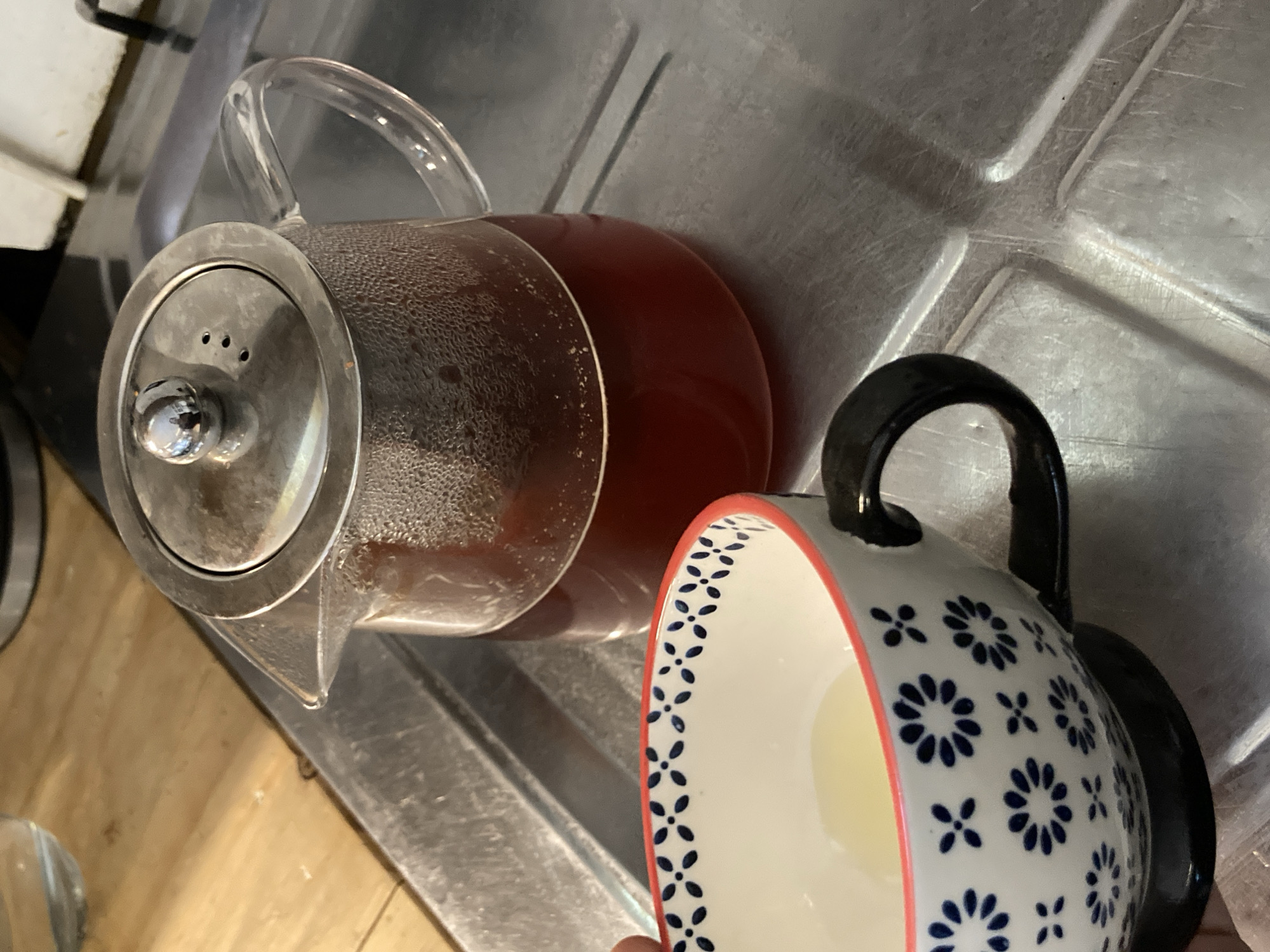 The image presents a close-up, tilted view of a clear glass teapot filled with a dark red liquid and a ceramic mug, both resting on a metallic surface. The teapot, featuring a clear glass handle and body, exhibits visible condensation on its inner walls, suggesting the liquid inside is hot. Its lid is made of a silver-toned metal, equipped with a central knob and three small circular holes, likely for straining or steam ventilation.
To the right of the teapot is a white ceramic mug, partially held by a hand that is barely visible at the bottom right. The mug has a prominent black handle and a vibrant red rim. Its exterior is decorated with a charming pattern of small blue four-petal floral shapes alternating with larger blue multi-petal flowers.
The setting appears to be an indoor space, possibly a kitchen, with the items placed on a brushed metallic surface that features raised ridges and troughs, characteristic of a sink's draining board. A light-colored wooden surface is visible to the left, suggesting a countertop. The overall scene captures a moment related to preparing or serving a hot beverage. No text is visible in the image.
