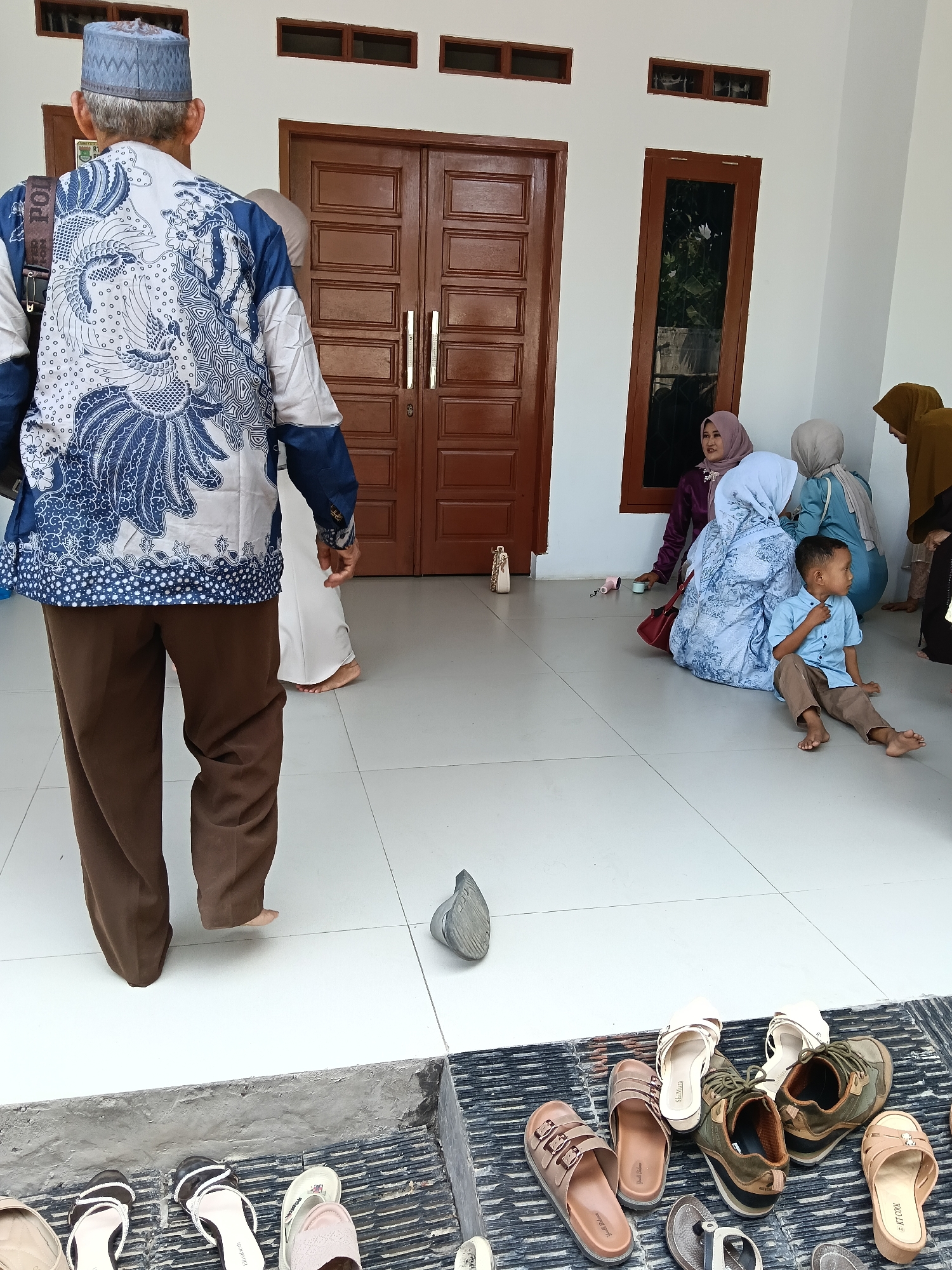 The image captures an outdoor scene in Tegal Angus, Indonesia, where a group of people are gathered on a tiled patio in front of a building with brown double doors and windows. In the foreground, an elderly man, viewed from the back, wears a patterned blue and white batik shirt and brown trousers, along with a light blue cap. To his right, a young boy in a light blue shirt and brown pants sits on the ground. Several women in Islamic attire, including hijabs, are also seated on the patio. Various footwear, including sandals and shoes, are arranged neatly on a mat near the steps leading down from the patio. The environment appears to be a residential or communal area, possibly in front of a mosque or a private residence.