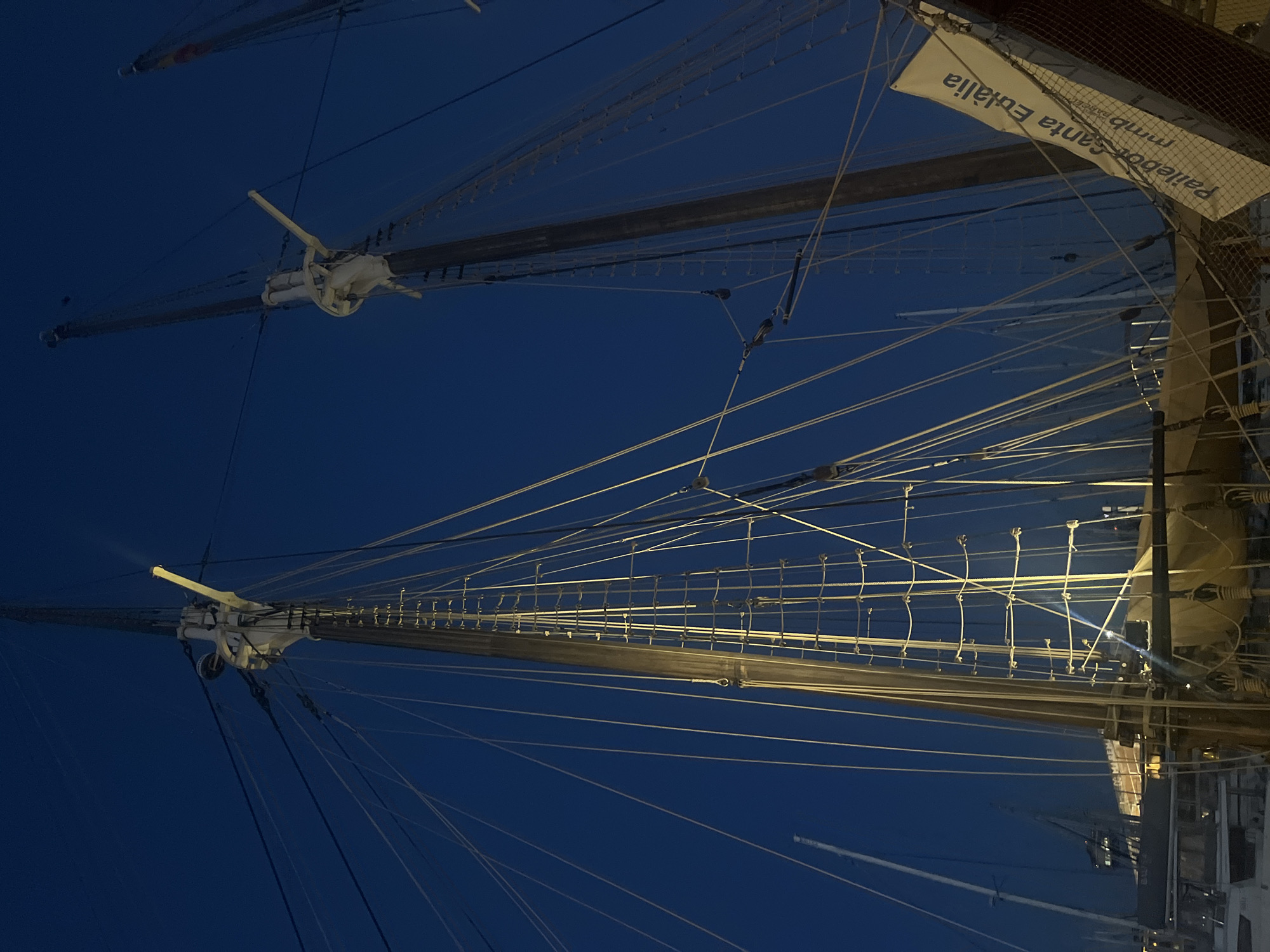 The image depicts the rigging of a tall ship against a dark blue sky. Two masts are prominently featured, with numerous ropes and lines extending from them. A banner with the text "Pallebot Santa Eulàlia" is visible on the right side of the image. The location is likely the port area of Barcelona, Spain, where the Santa Eulàlia, a historic schooner, is often docked. The scene suggests either early morning or late evening due to the low ambient light.