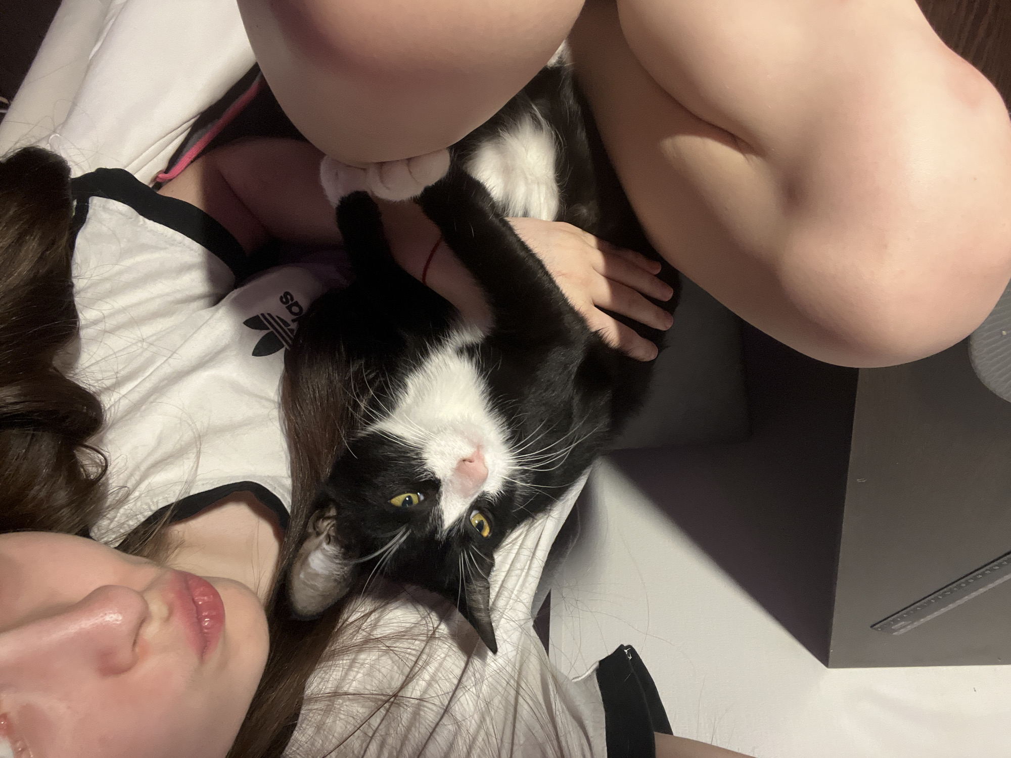 A woman lies on her back, knees bent, with a black and white cat resting on her stomach. Her right hand rests on the cat. She is wearing a white Adidas t-shirt. The location is not identifiable as Saint Petersburg, Russia.