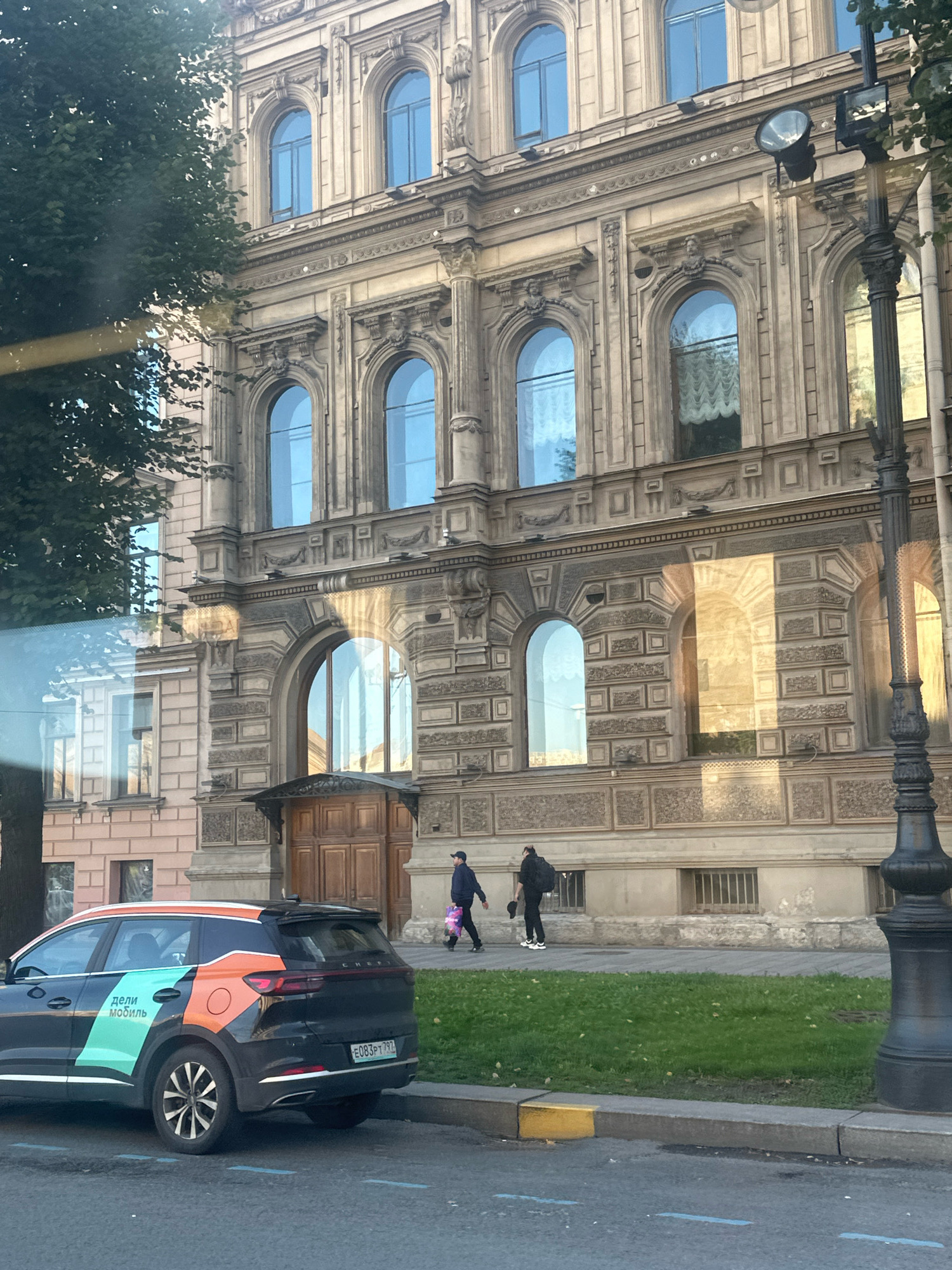 The image shows a street scene in Saint Petersburg, Russia, with a large, ornate building as the backdrop. The building features classical architecture with arched windows, decorative carvings, and a light-colored facade. Sunlight casts strong shadows across the building's surface.
In the foreground, a dark-colored car with colorful decals is parked on the street. The decals include text in Russian, "дели мобил," which translates to "taxi mobile" or "car sharing." The car has a license plate that reads "E083PT 797."
Two individuals are walking on the sidewalk in front of the building. One is wearing a dark jacket and pants, carrying a bag. The other is wearing a dark hoodie and pants, carrying a backpack and a hat. A black lamppost with an elaborate design stands on the right side of the image. The street in front of the car has blue painted lines. A strip of green grass is visible between the sidewalk and the street.