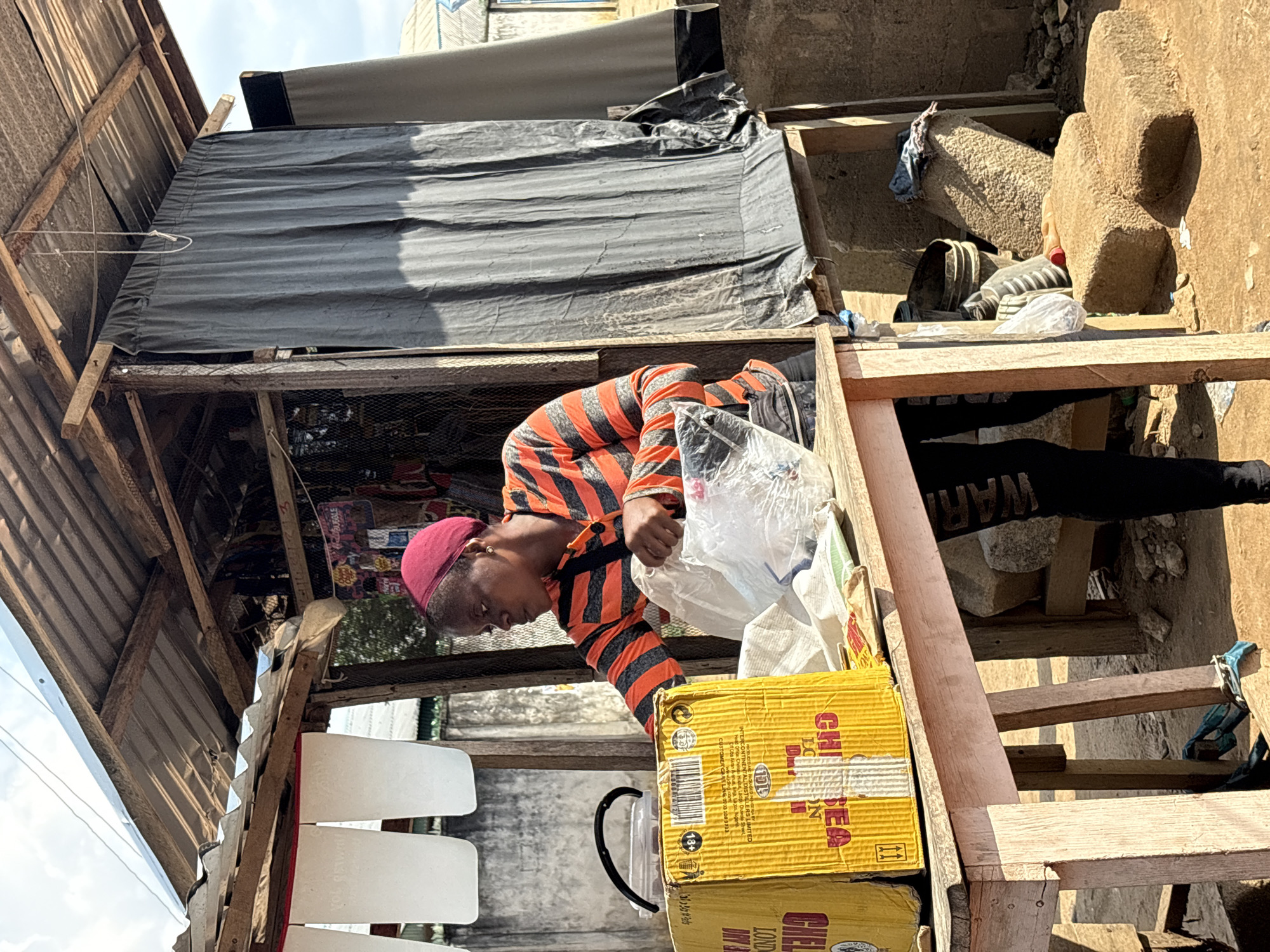 A person wearing a striped orange and black shirt and a maroon cap is reaching into a plastic bag held over yellow boxes labeled "CHELSEA LONDON DRY GIN" inside a small, rudimentary wooden structure. White, rectangular objects hang nearby. The structure has a corrugated metal roof and is covered with a gray tarp. The area appears to be Ikot Ekpene, Nigeria, based on the product label. Nearby are stones and various objects, suggesting a cluttered environment.