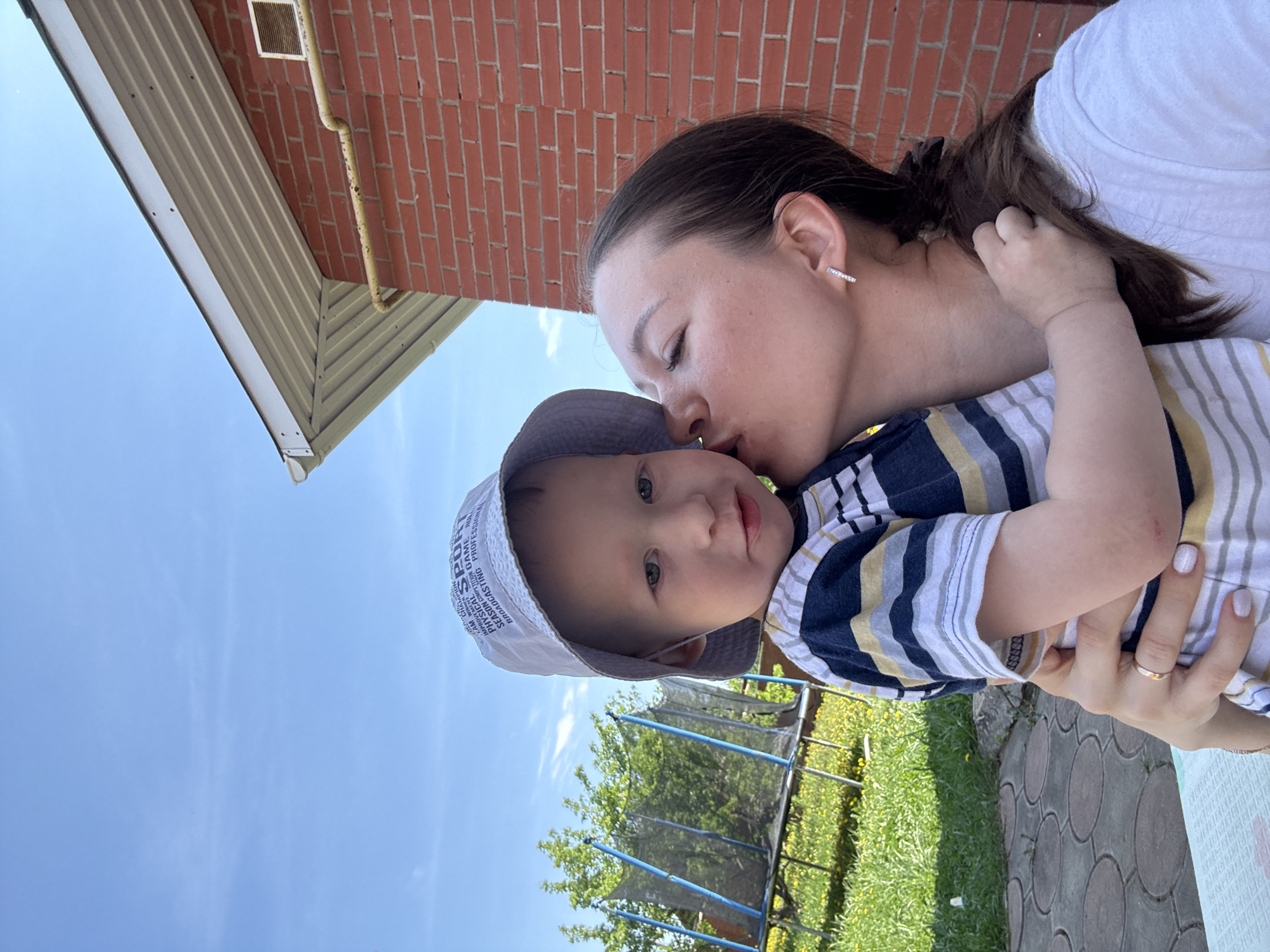 A woman is holding and kissing a young child. The woman has dark hair pulled back and is wearing a white shirt. The child is wearing a hat and a striped shirt. They are outdoors in a backyard. A brick building and a trampoline are visible in the background. The location is Ферма, Russia.