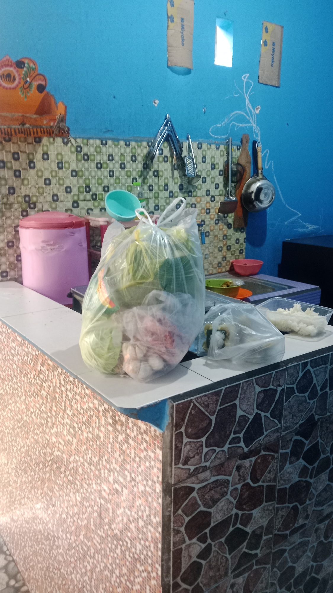 The image shows a domestic kitchen scene, possibly in Denpasar, Indonesia, characterized by vibrant blue walls and a patterned tile backsplash. A large plastic bag filled with fresh produce, including what appears to be cabbage, garlic, and possibly onions, is the central focus, placed on a tiled countertop. To the left of the produce bag, a pink cylindrical container with a lid sits near the edge of the counter. The background reveals a sink area with a faucet and several utensils hanging on the wall, including a rolling pin and a frying pan. Two small, rectangular pieces of cardboard, possibly repurposed as signage or decoration, are attached to the blue wall. The countertop itself is tiled with a mosaic of light-colored squares and darker, irregular stone-like shapes. The overall impression is one of a functional, lived-in kitchen space.