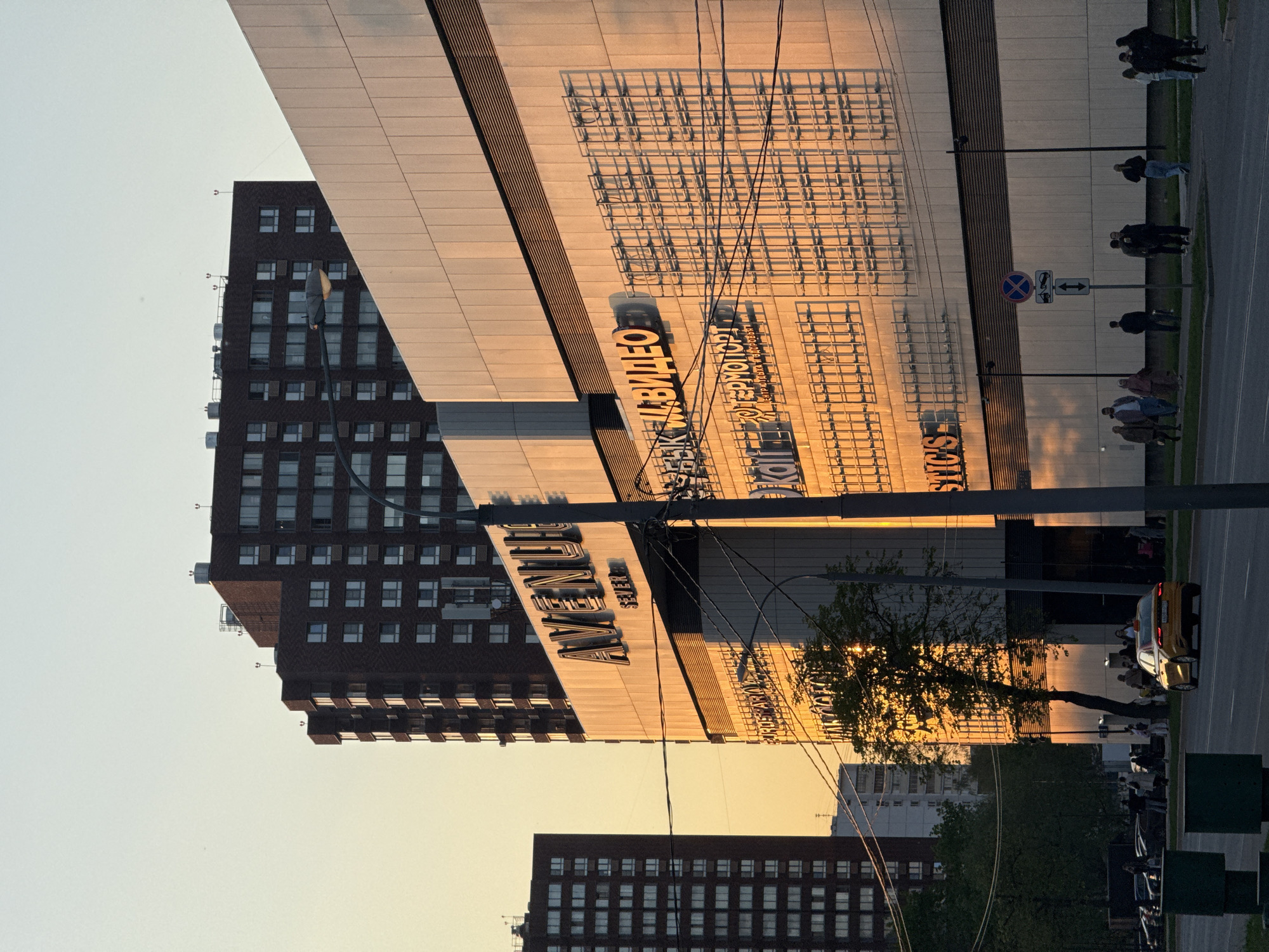 The image captures a street scene in Moscow, Russia, during what appears to be the late afternoon or early evening, given the warm, golden light. The foreground is dominated by a multi-story building with modern architecture, featuring a combination of dark brick and light-colored panels. Prominently displayed on the building is the word "AVENUE". Another smaller building of brick and windows is seen in the background. The sky is partially visible, transitioning from a light blue to a yellow hue. On the sidewalk, pedestrians can be seen walking and standing. In the street, cars are parked along the curb. Overhead, power lines crisscross the scene, adding complexity to the cityscape.
