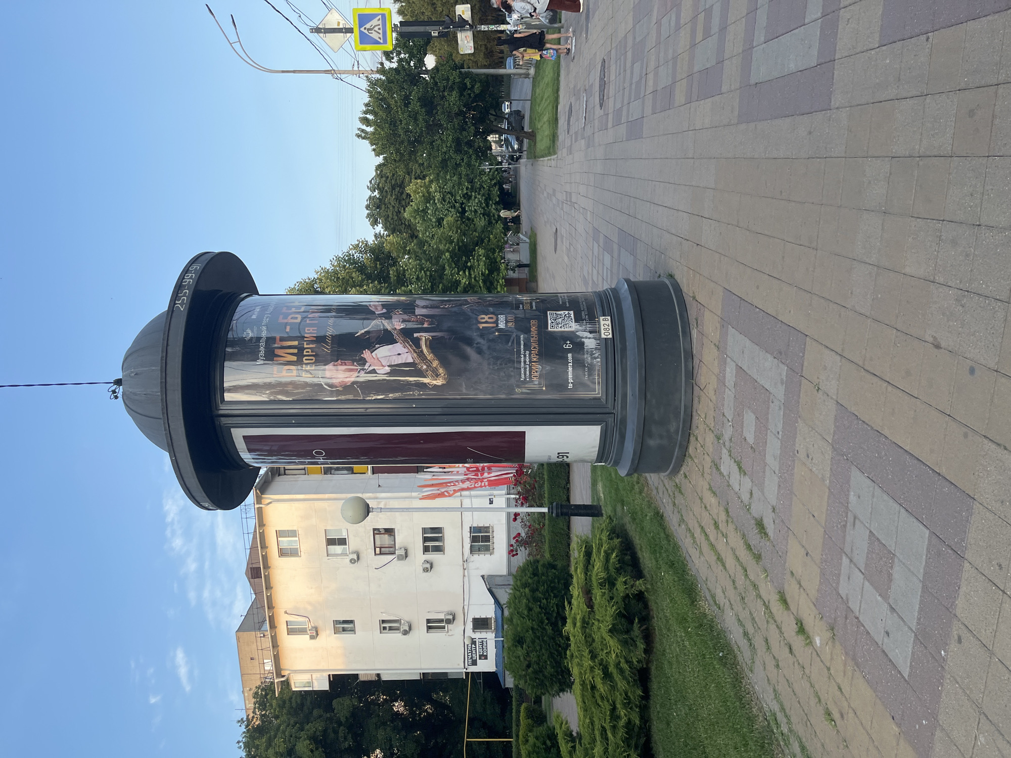 The image depicts a street scene in Krasnodar, Russia, on a bright day with a clear blue sky and scattered clouds. In the foreground, a cylindrical advertisement pillar stands prominently on a paved sidewalk. The pillar displays a poster advertising a jazz concert, featuring a saxophone player and the text "Биг-Бен Георгия Гай" and "Юрий Красильников". A phone number "255-99-91" is also visible on the pillar.
To the left of the pillar, a well-maintained green lawn and manicured bushes are visible, bordering a white multi-story building. The building has multiple windows and air conditioning units. A street lamp stands between the pillar and the building.
The sidewalk is made of interlocking paving stones in various shades of brown, yellow, and grey, with some grass growing through the cracks. Further down the street, a pedestrian crossing with a yellow and blue sign is present, along with a traffic light. Several people are walking on the sidewalk in the middle ground and background. Trees line the right side of the street, with a road and cars visible beyond them. The overall environment suggests a typical urban setting with residential and commercial elements.