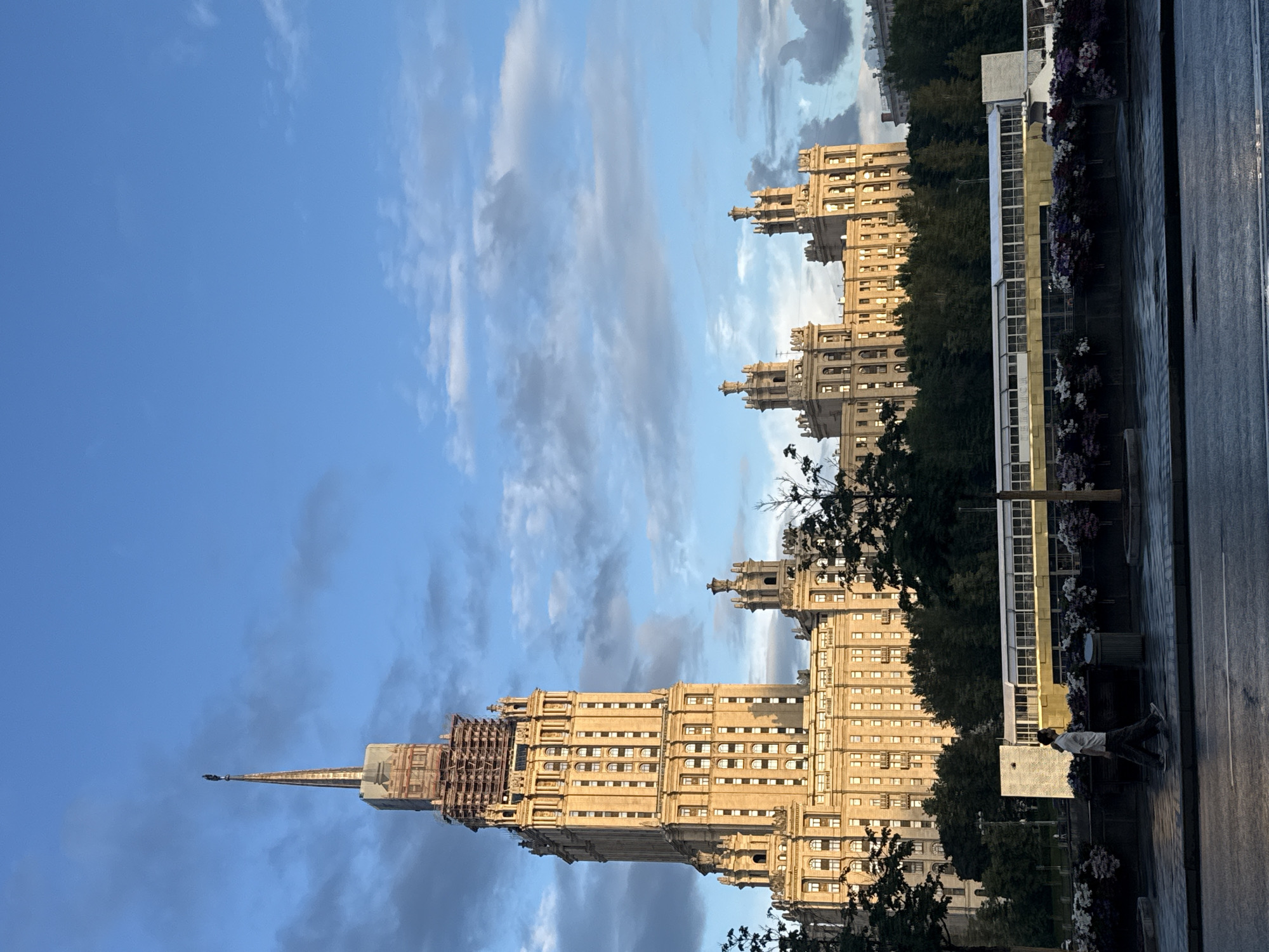 The image shows a wide shot of the Stalinist-era skyscraper, the Kudrinskaya Square Building, in Moscow, Russia. The building, with its distinctive spire, is bathed in warm, late afternoon sunlight, casting long shadows. The sky above is a vibrant blue, with scattered, dramatic clouds. In the foreground, a man walks along a paved walkway, passing a row of planters filled with colorful flowers. A modern-looking structure, possibly a pavilion or a transit stop, runs horizontally across the mid-ground, partially obscured by trees. The overall impression is one of urban grandeur under a dynamic sky.