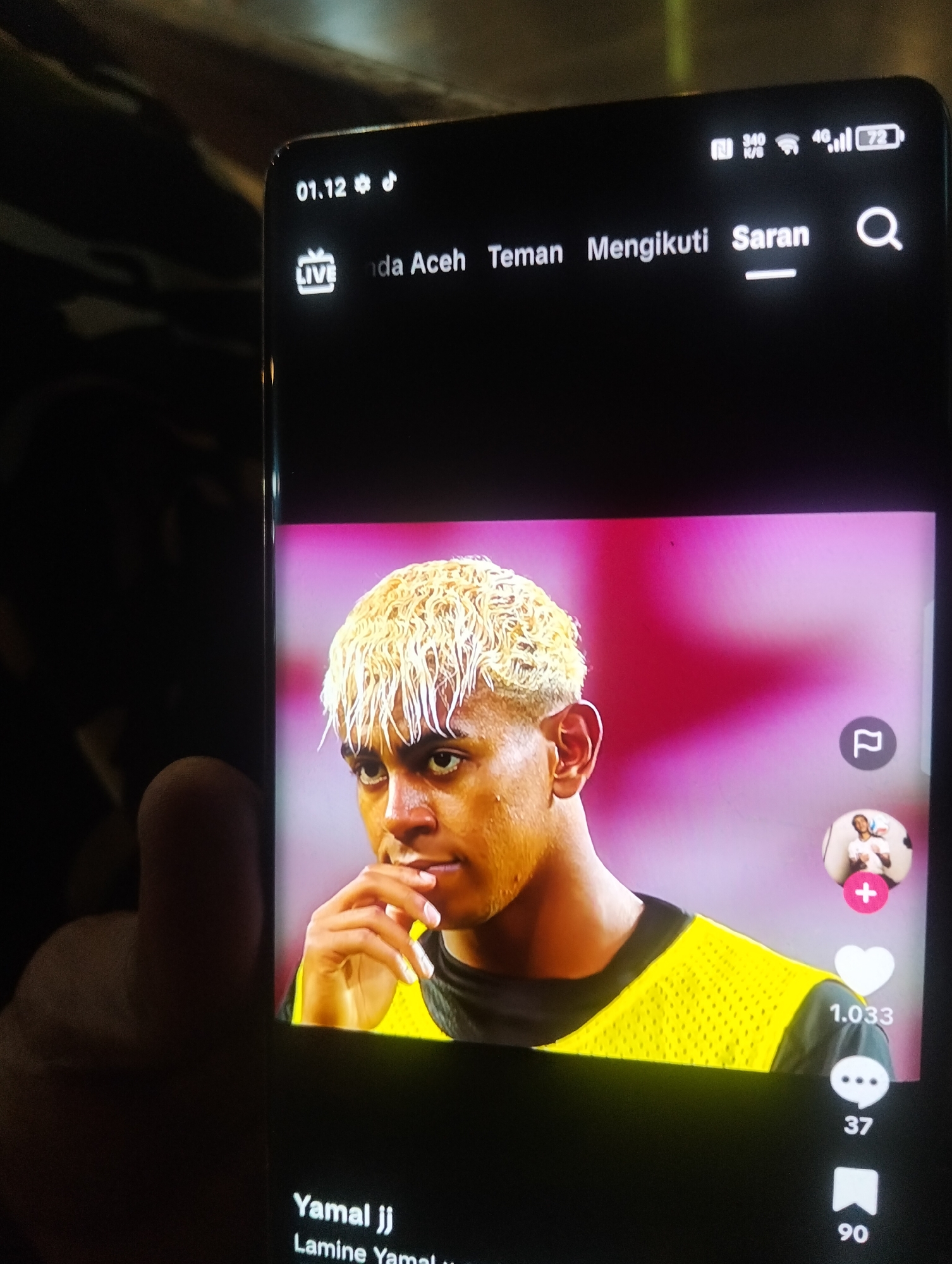 The image is a close-up of a smartphone screen displaying a social media video. The video features a person with bleached blonde hair and a yellow vest. On the screen, there are icons and text typical of a video-sharing platform, including a like count of "1.033", a comment count of "37", and a share count of "90". The top of the screen shows the date "01.12" and signal strength indicators. The bottom of the screen displays the username "Yamal jj" and "Lamine Yamal". The background of the video is a blurred pink color. The surrounding environment appears to be dark, suggesting the photo was taken in low light conditions. There is no visual information in the image that places it in the city of Payagaboh, Indonesia.