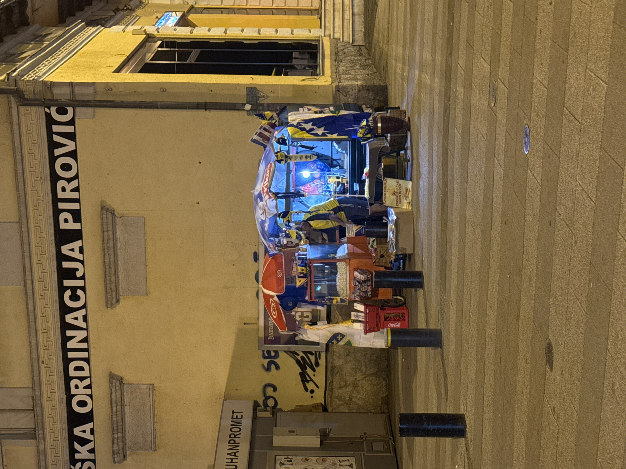 A street scene at night in Ilidža, Bosnia and Herzegovina, shows a food stand selling corn in a cup. The stand is set up in front of a building with a yellow facade. A person, wearing a yellow vest and dark pants, is standing behind the stand. The stand is decorated with Bosnian flags and has a red umbrella providing shade. There is a popcorn machine and other items displayed for sale. The ground is paved with stone tiles.