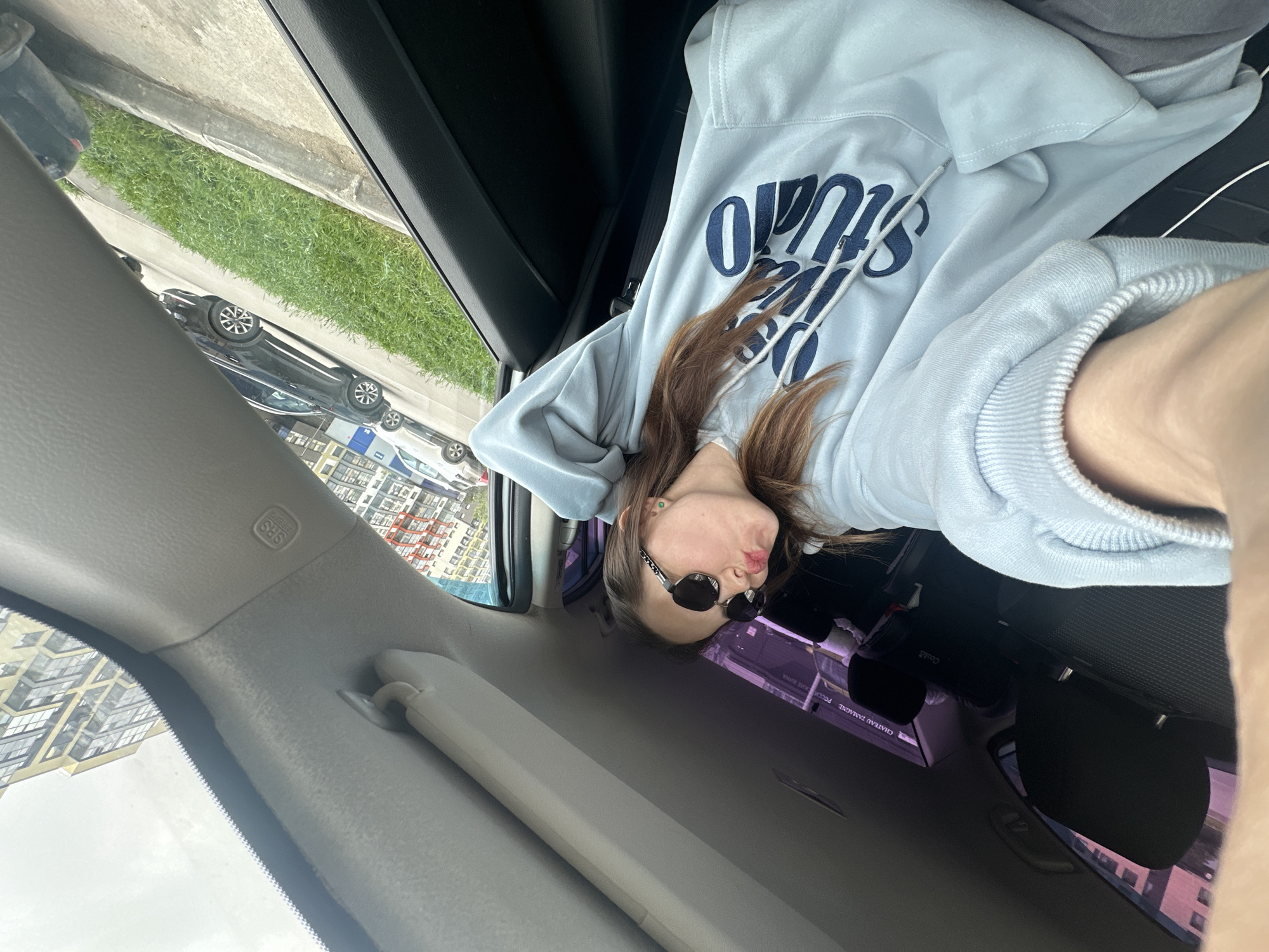 A person in a light blue hooded sweatshirt and sunglasses is seated in the passenger seat of a car. The car appears to be stationary. The interior of the car is visible, including the dashboard, door panel, and seating. Outside the window, a cityscape can be seen, with buildings and parked cars visible. Based on the Cyrillic text visible outside the window, this image was taken in Perm, Russia.