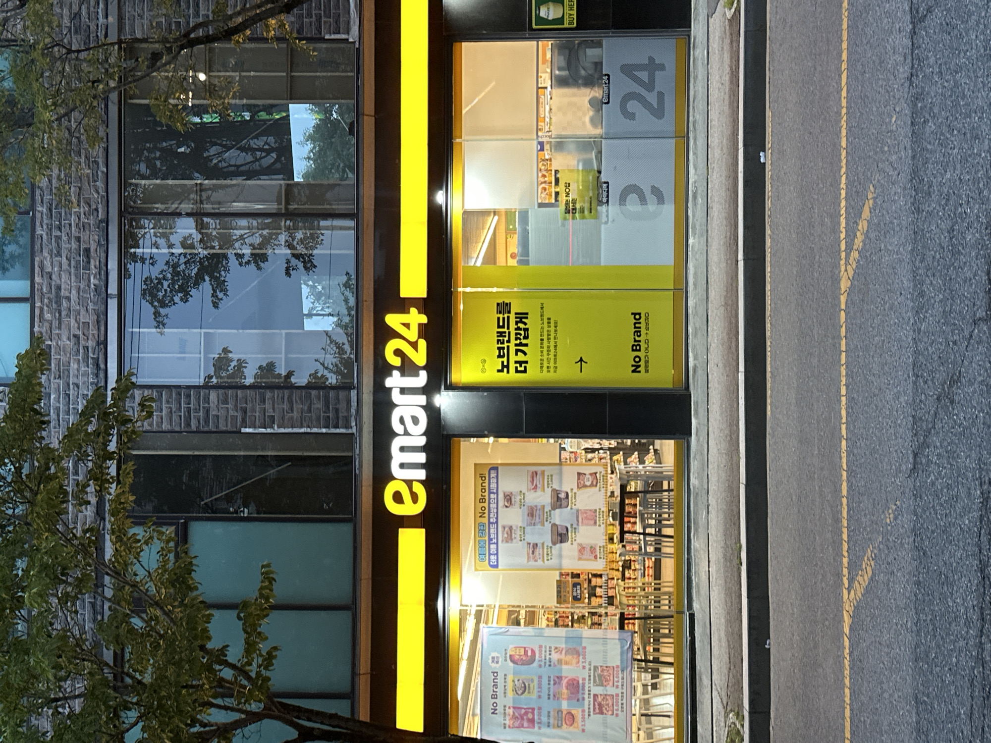 An exterior, eye-level, medium shot captures an E-mart 24 convenience store at dusk on a city street. The store's facade is illuminated by bright yellow signage reading "e-mart24" in large, bold lettering. Below the signage, the storefront features large glass windows offering a view into the well-lit interior. Promotional posters are displayed on the windows. One poster on the left, in Korean and English, advertises "No Brand" products with various item prices listed. A second poster, also in Korean, highlights "No Brand" products and encourages customers to visit. On the right side of the store, large numbers "24" are displayed, part of the store's branding. The store is situated on a paved street with yellow lane markings. Trees with green foliage are visible on either side of the building, their branches partially obscuring the upper floors. The overall impression is of a typical urban convenience store environment. The image was taken in Jeonju-si, South Korea.